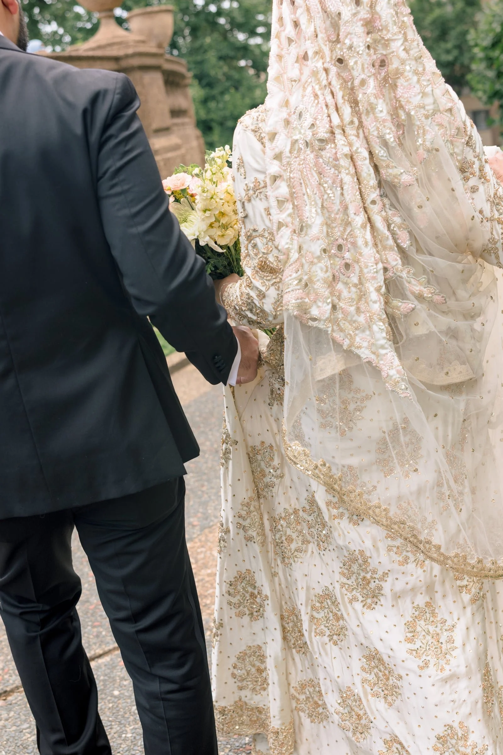 A bride and groom holding hands at their wedding ceremony outdoors, with the bride wearing a detailed, embellished white gown and the groom wearing a black suit, with a bouquet of flowers in the bride's hand.