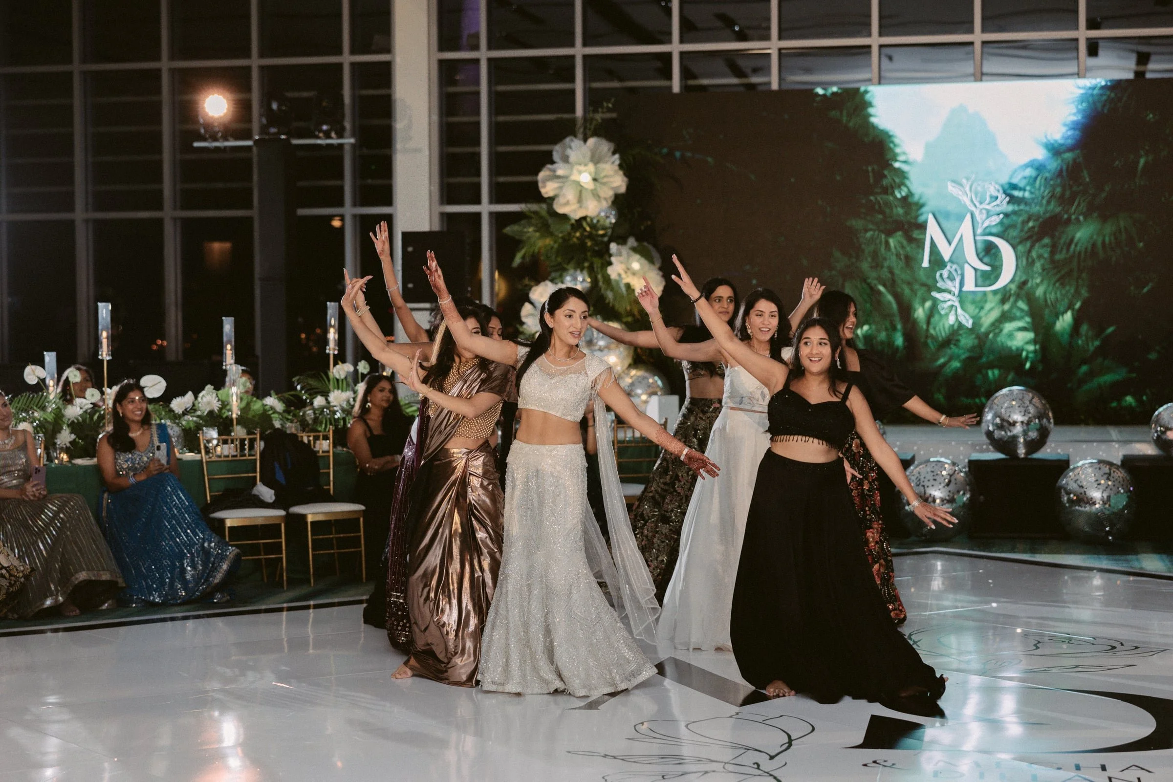 Group of women dancing at a formal event inside a venue with large glass windows and illuminated decorations, with some women seated at tables watching the performance.