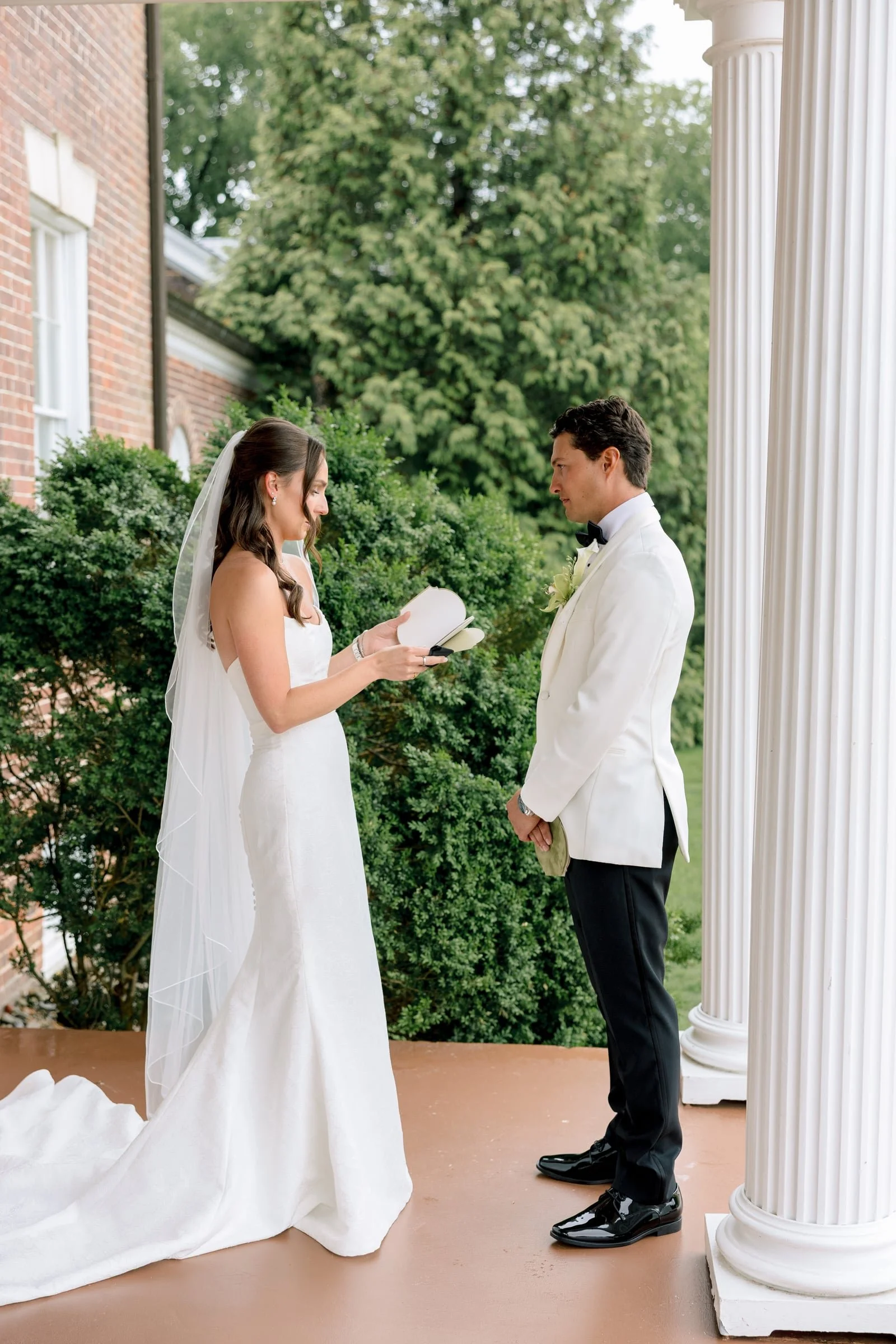 A bride and groom are standing outdoors on a porch with white columns, exchanging vows. The bride is wearing a white wedding gown with a veil, and the groom is dressed in a white tuxedo with a black bow tie. There is greenery and a brick building in 