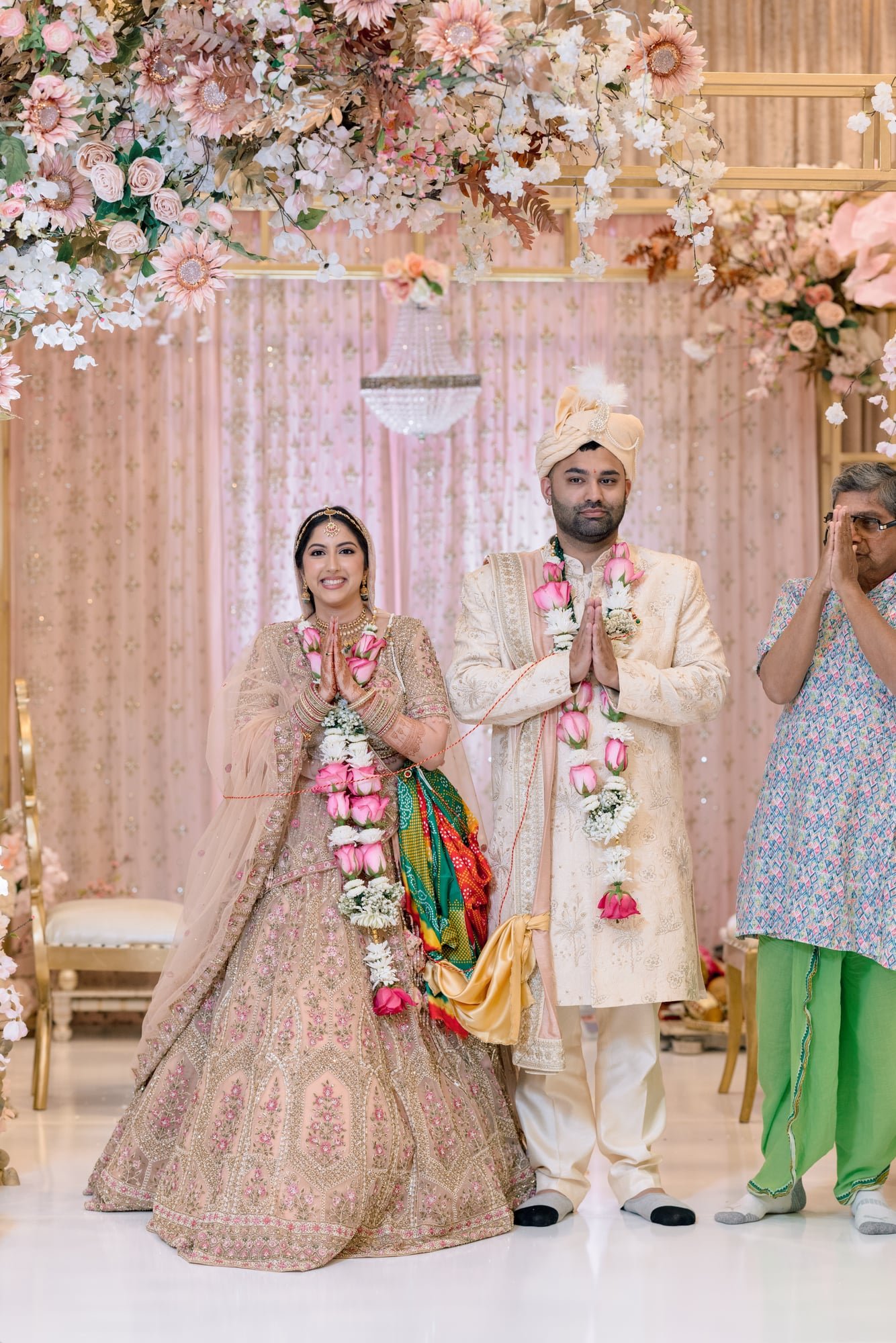 An Indian wedding ceremony with a bride and groom dressed in traditional attire, standing under a floral canopy with pink and white flowers, a chandelier in the background, and a person on the right side covering their face with their hands.