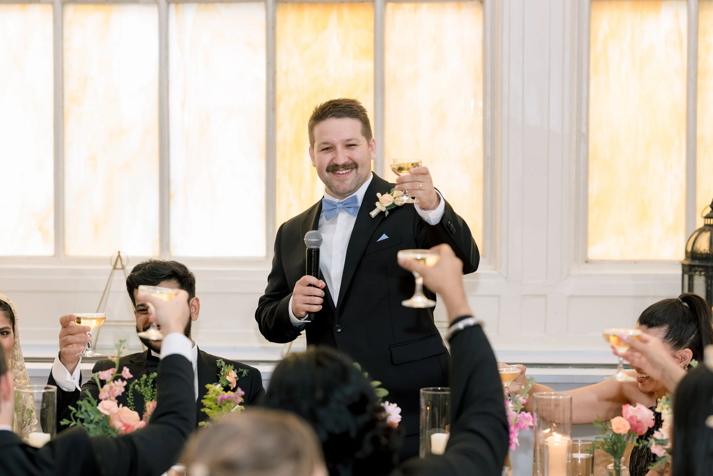 A man in a suit and bowtie holding a microphone and making a toast at a wedding reception, surrounded by guests raising glasses.
