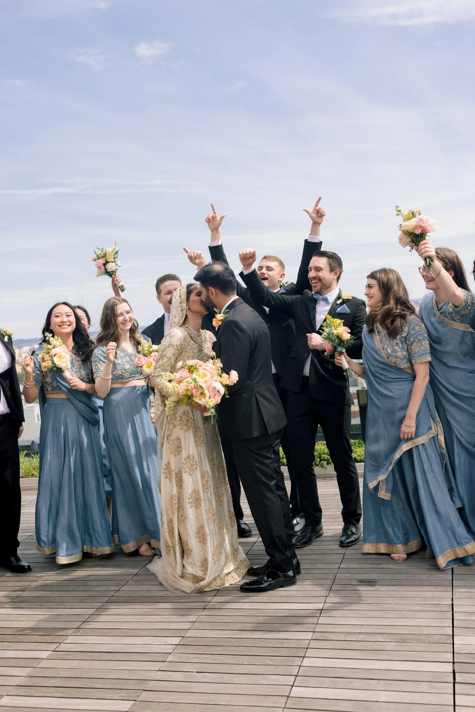A group of people at a wedding celebration outdoors, with the bride and groom kissing in the center. The bride wears an ornate traditional dress, and the groom is in a black tuxedo. Several women in blue dresses and men in suits are celebrating, smil
