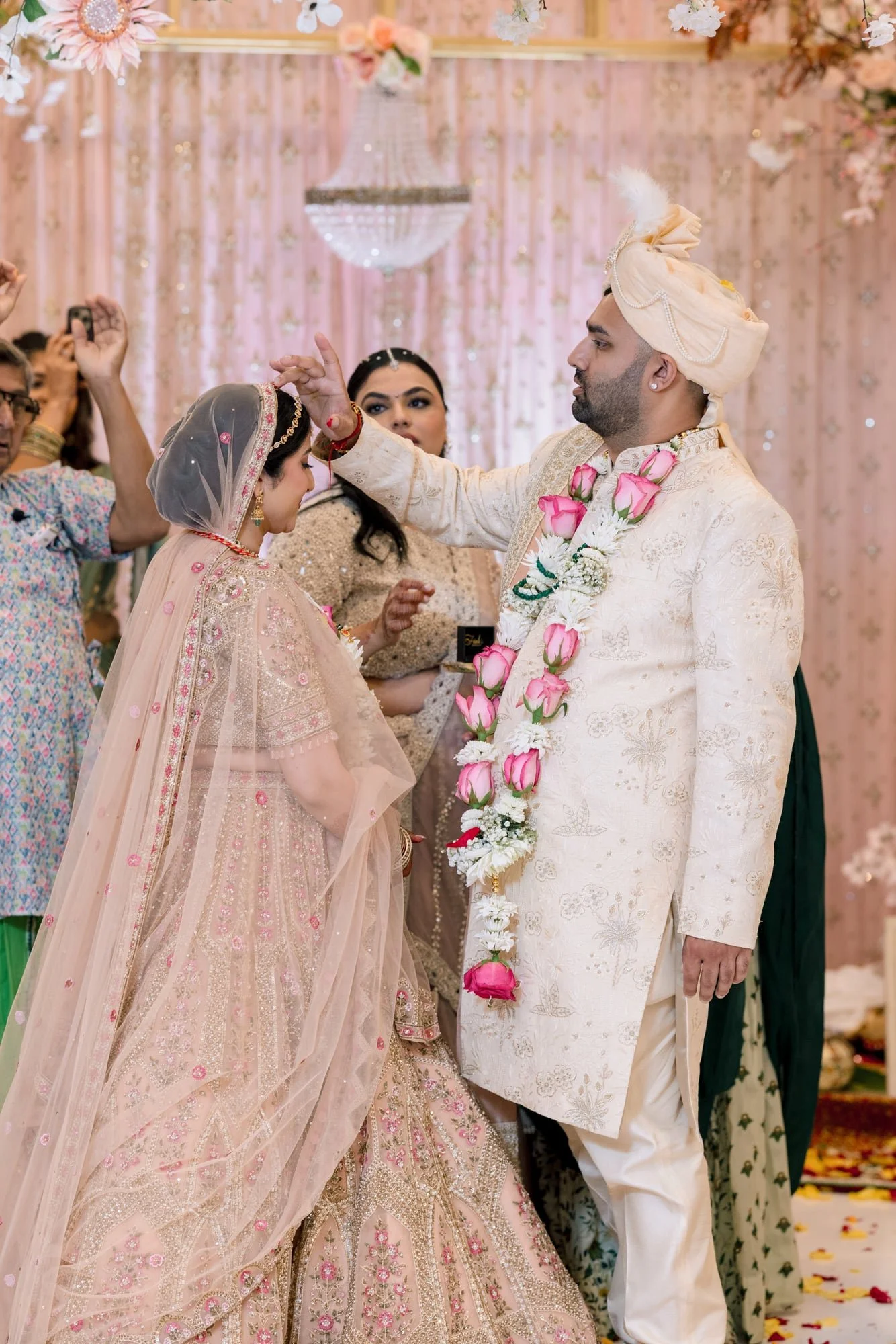 Indian wedding ceremony with groom in ivory Sherwani and turban and bride in pink lehenga and veil, surrounded by family and friends, under pink floral decor, during the wedding rituals.