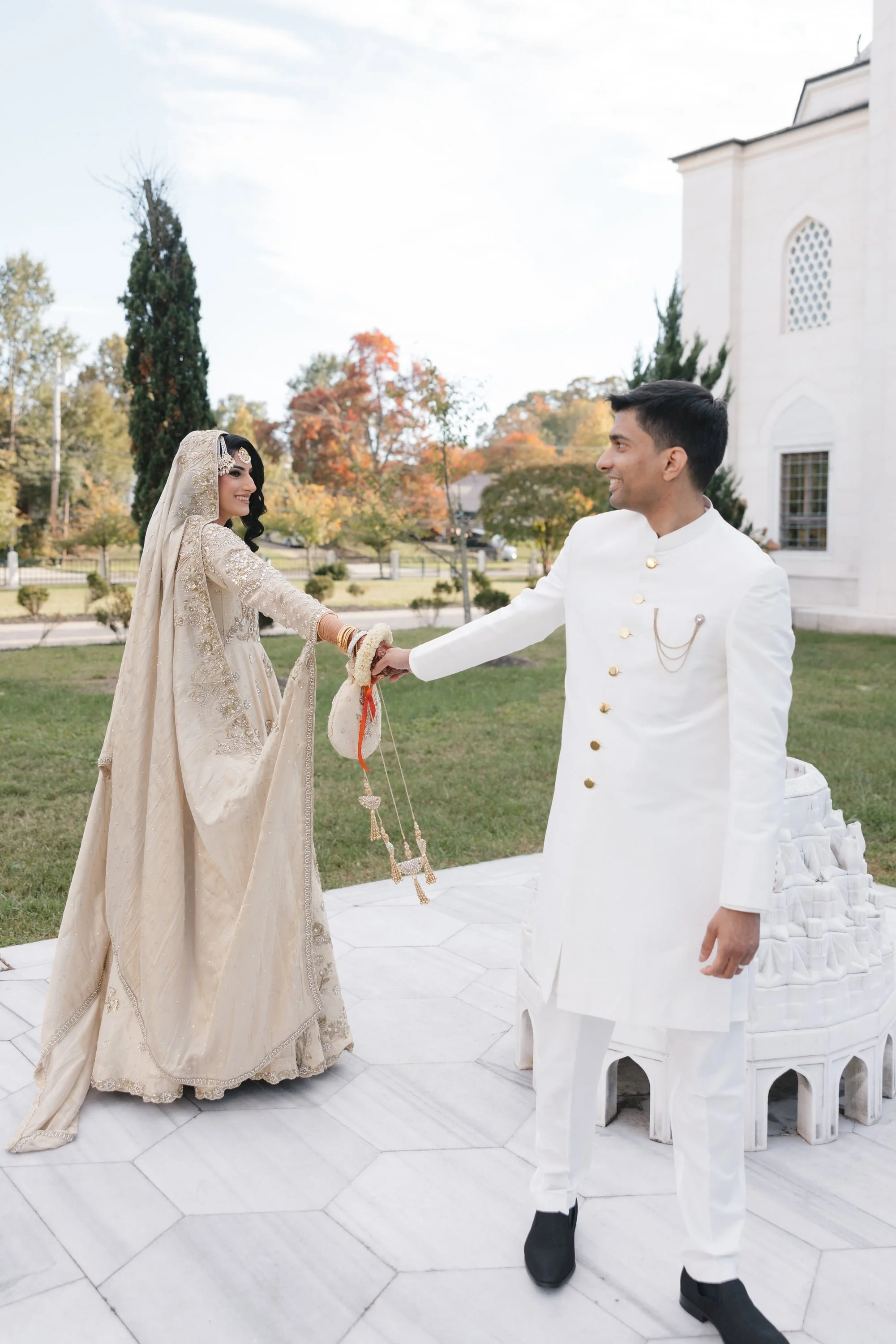 A bride and groom holding hands outdoors in front of a white building with trees displaying fall foliage in the background.
