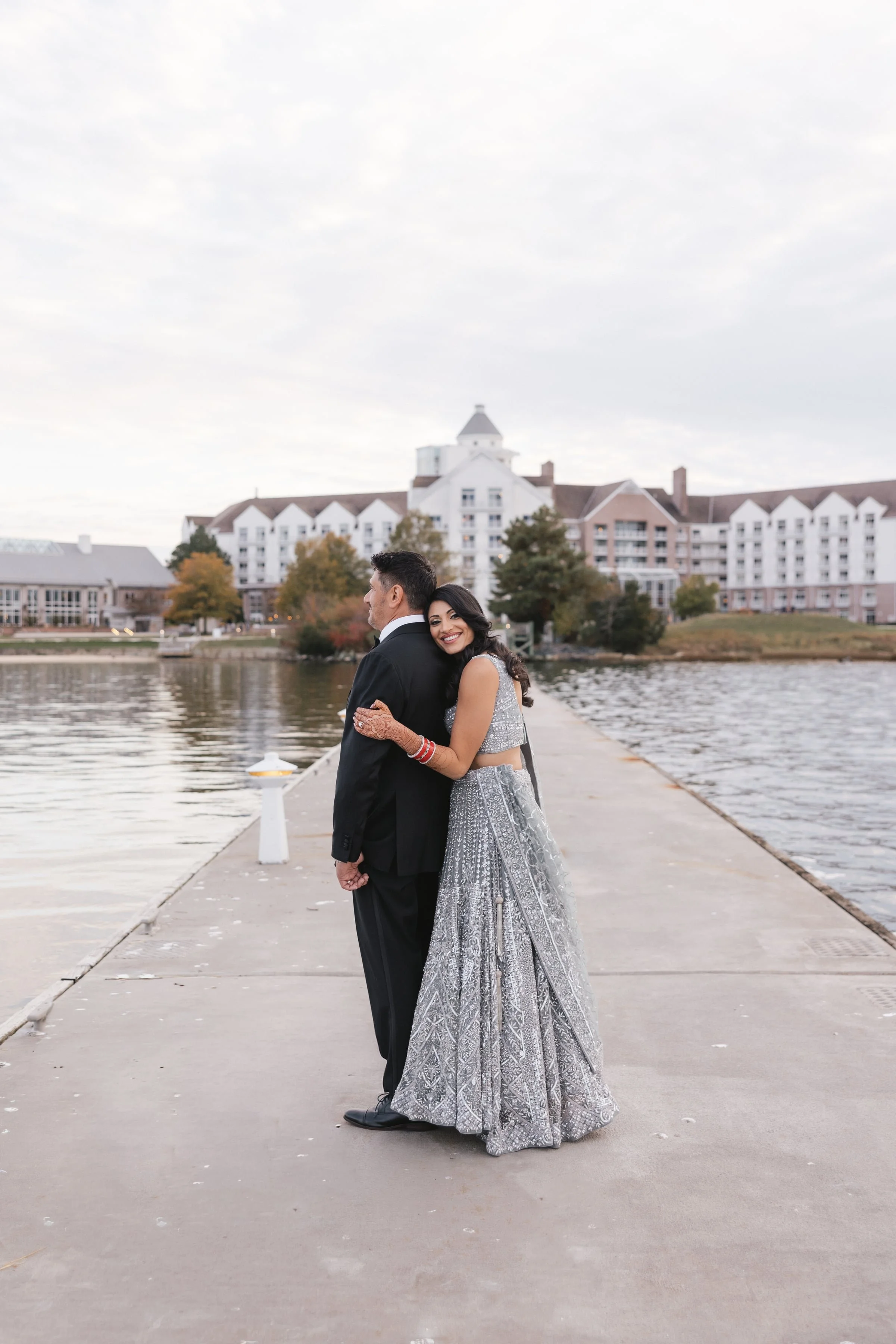 A bride and groom standing on a dock by a lake, with the bride smiling and leaning her head on the groom's shoulder, in front of a large hotel or resort building, during daytime with cloudy sky.