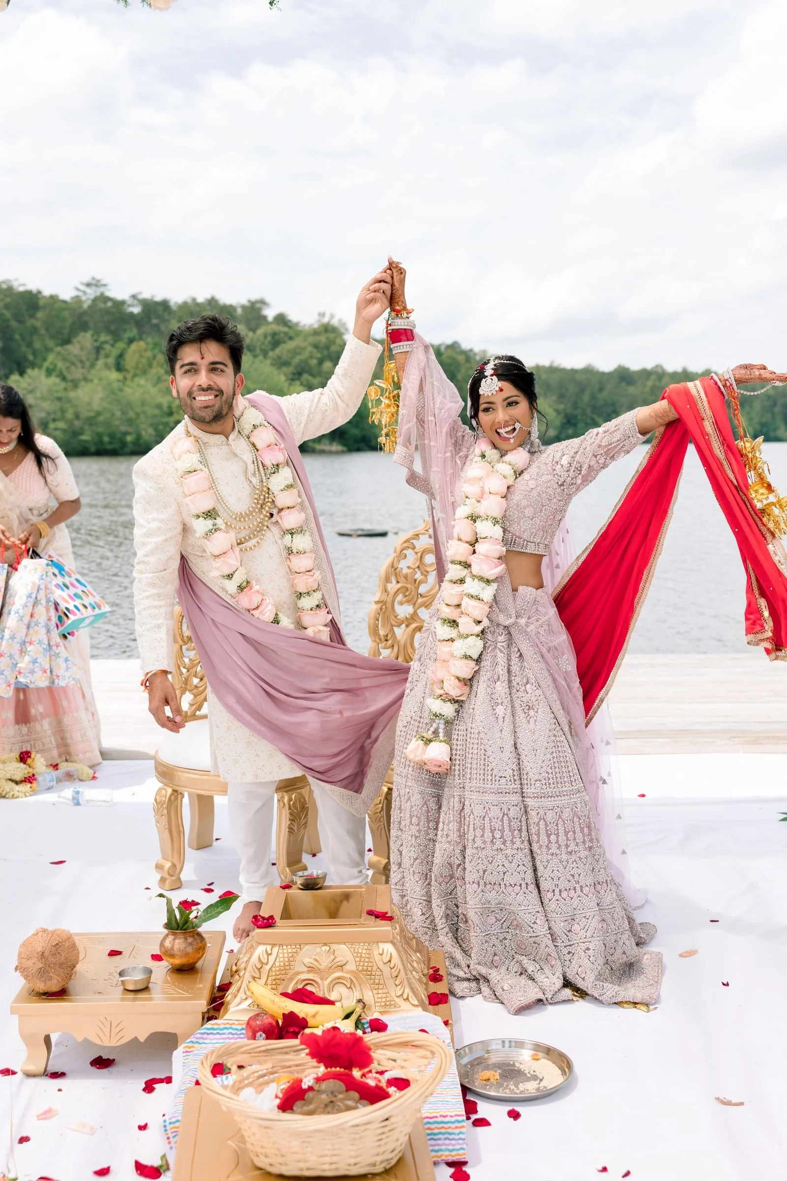 Indian bride and groom celebrating their wedding outdoors by a lake, holding hands with floral garlands, with wedding offerings and decorations around them.