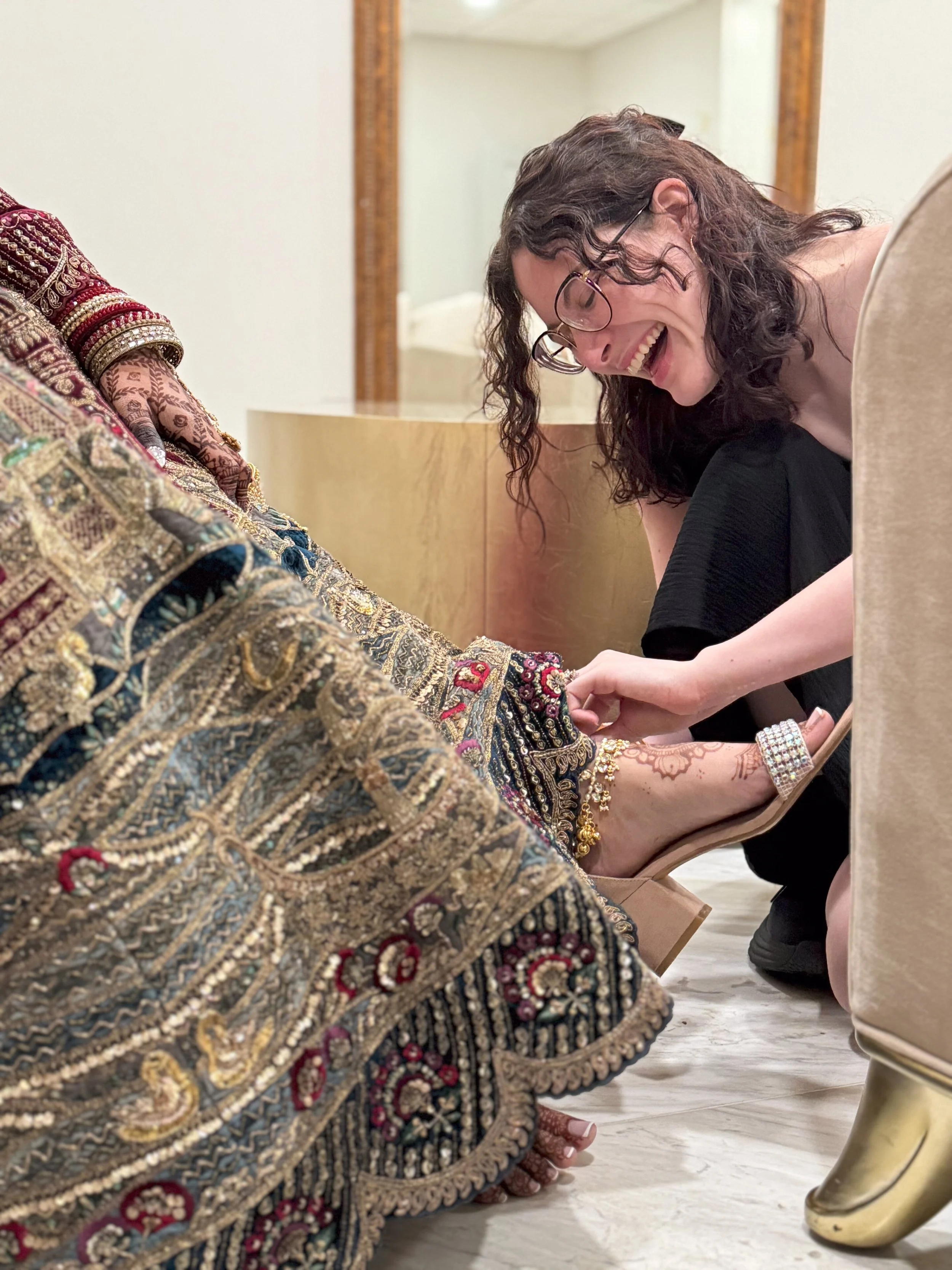 A woman kneeling on the floor, smiling and leaning forward to touch the hand of someone wearing ornate, embroidered clothing. The woman has curly hair, glasses, and is wearing a black top and tan pants. There are bangles on her wrist and a bracelet, and henna designs on her hand.