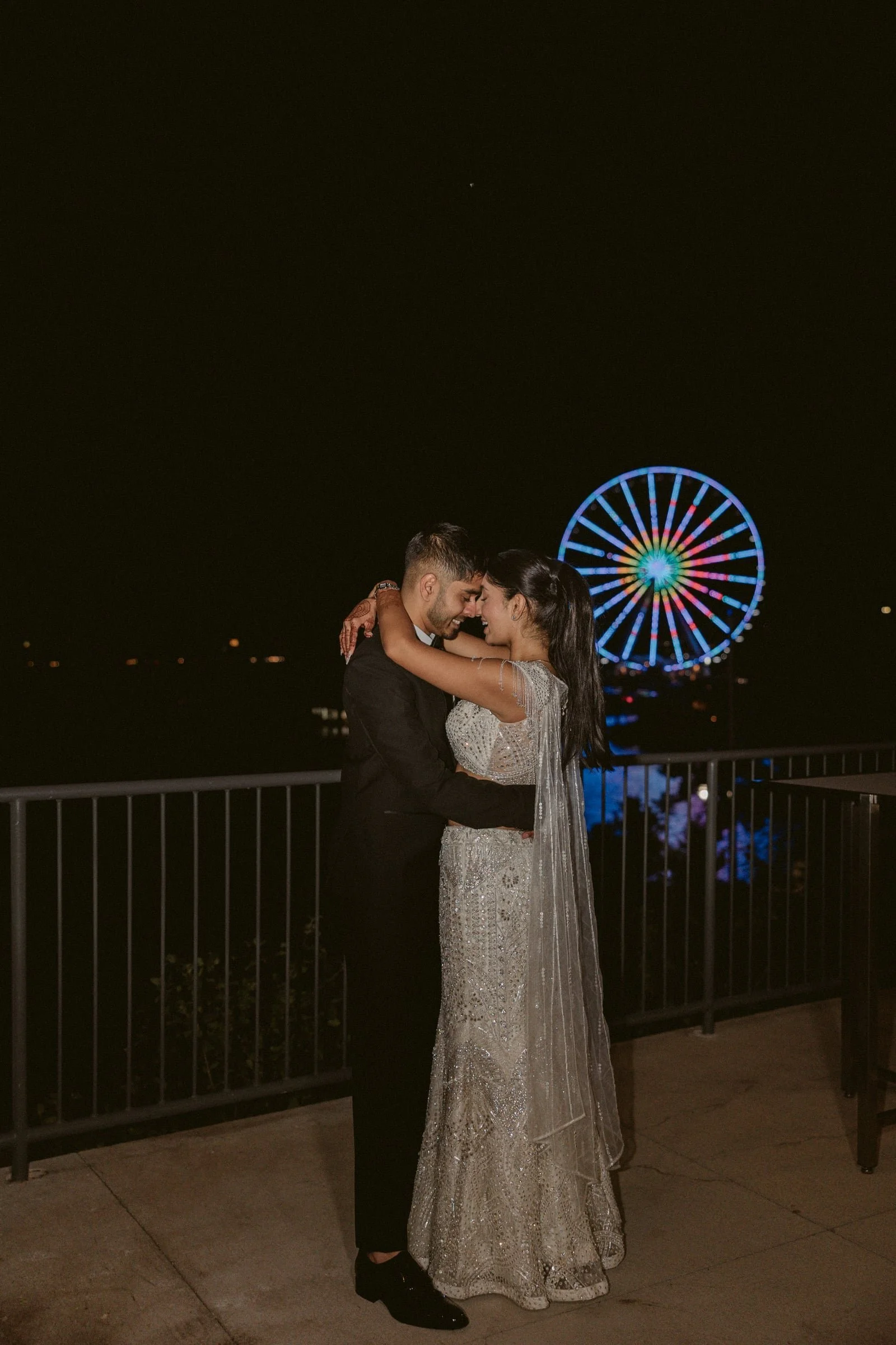 A couple dressed in formal attire embracing on a night-time amusement park pier with a brightly lit Ferris wheel in the background.