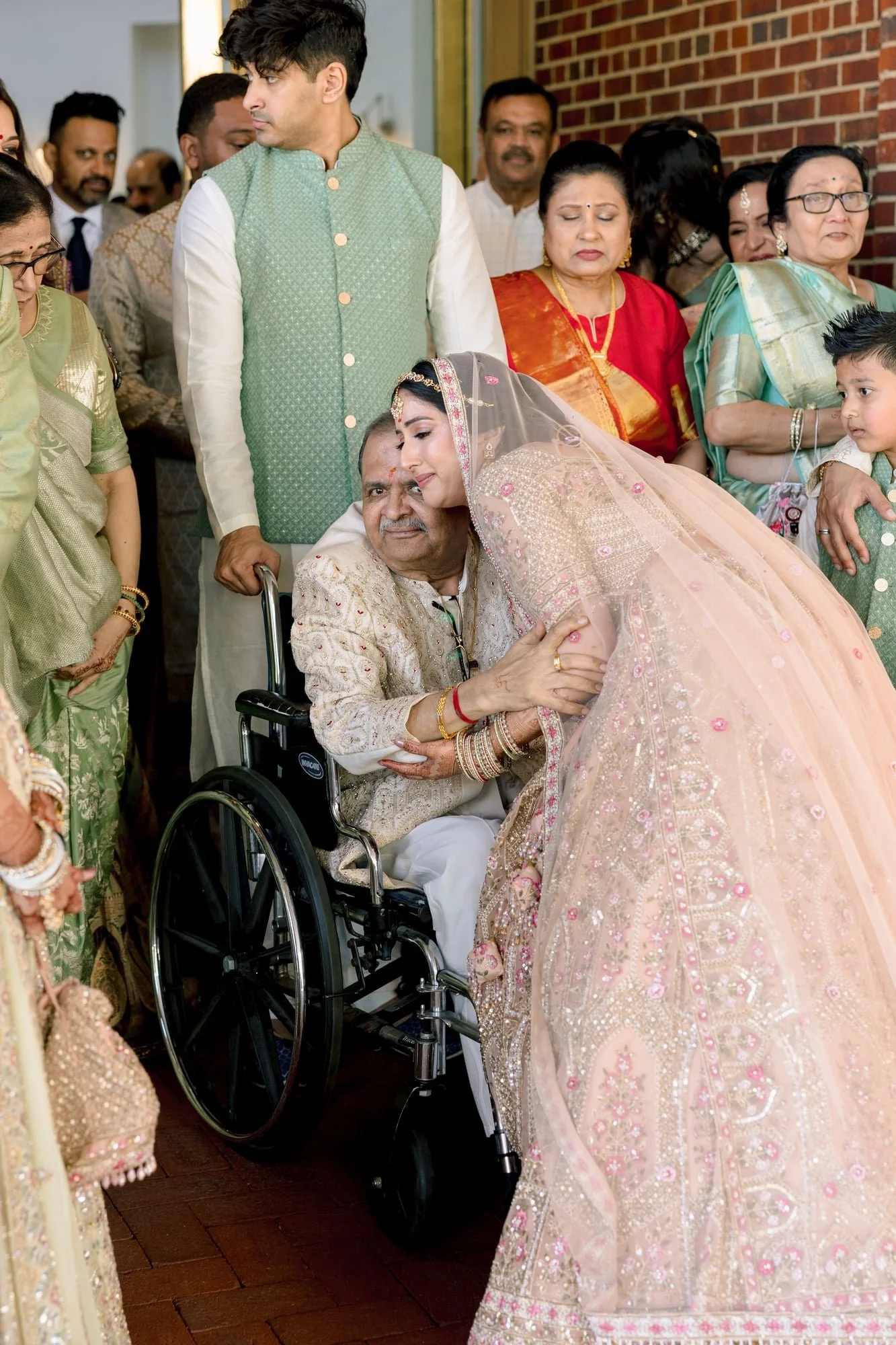 A Hindu wedding ceremony with a bride in a pink, gold, and silver embroidered traditional dress kissing an elderly man in a wheelchair, surrounded by family members dressed in colorful traditional attire.