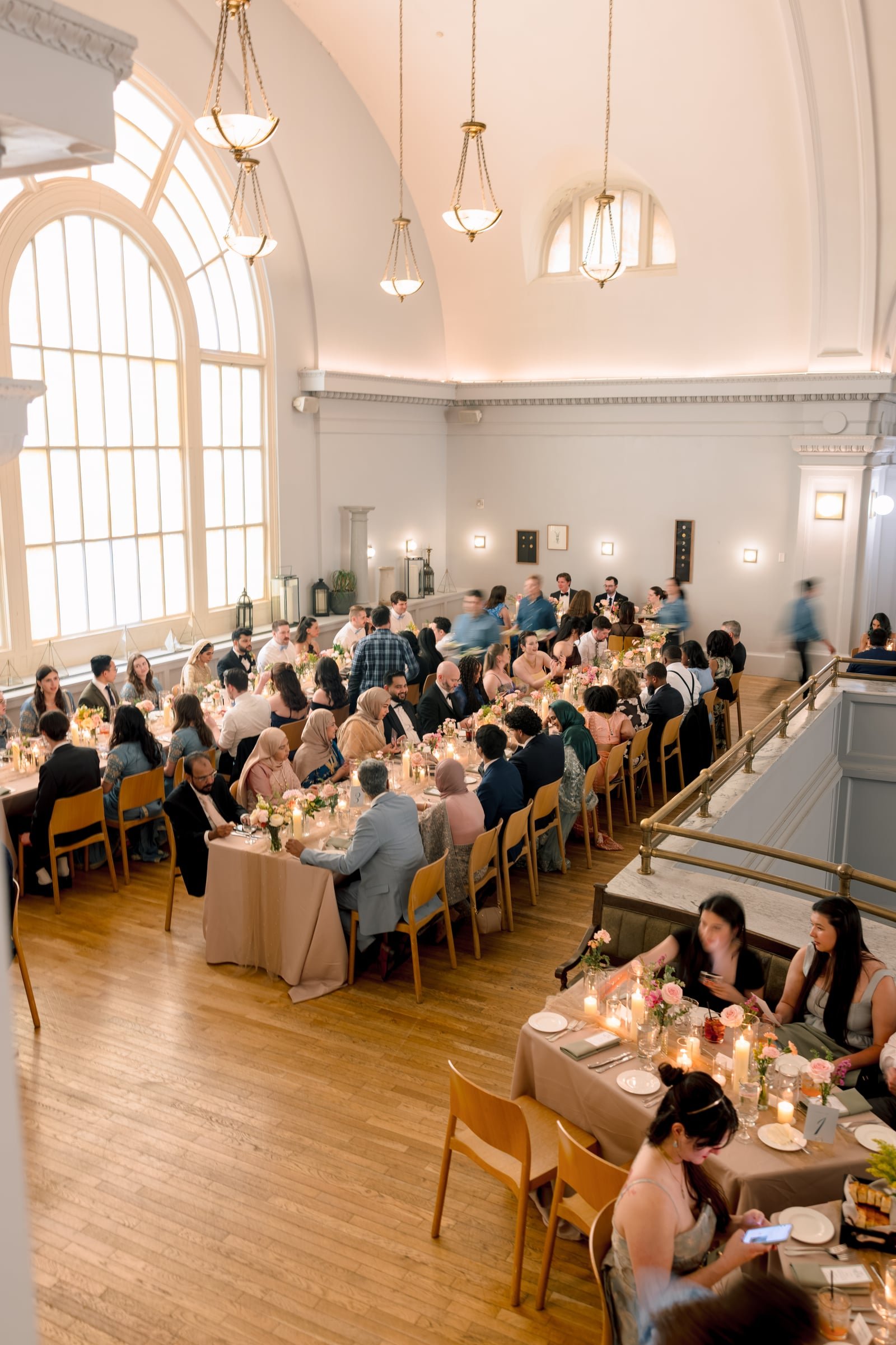 A large banquet hall filled with guests seated at long tables decorated with flowers and candles, with high arched windows, beige walls, and elegant lighting fixtures hanging from the ceiling.