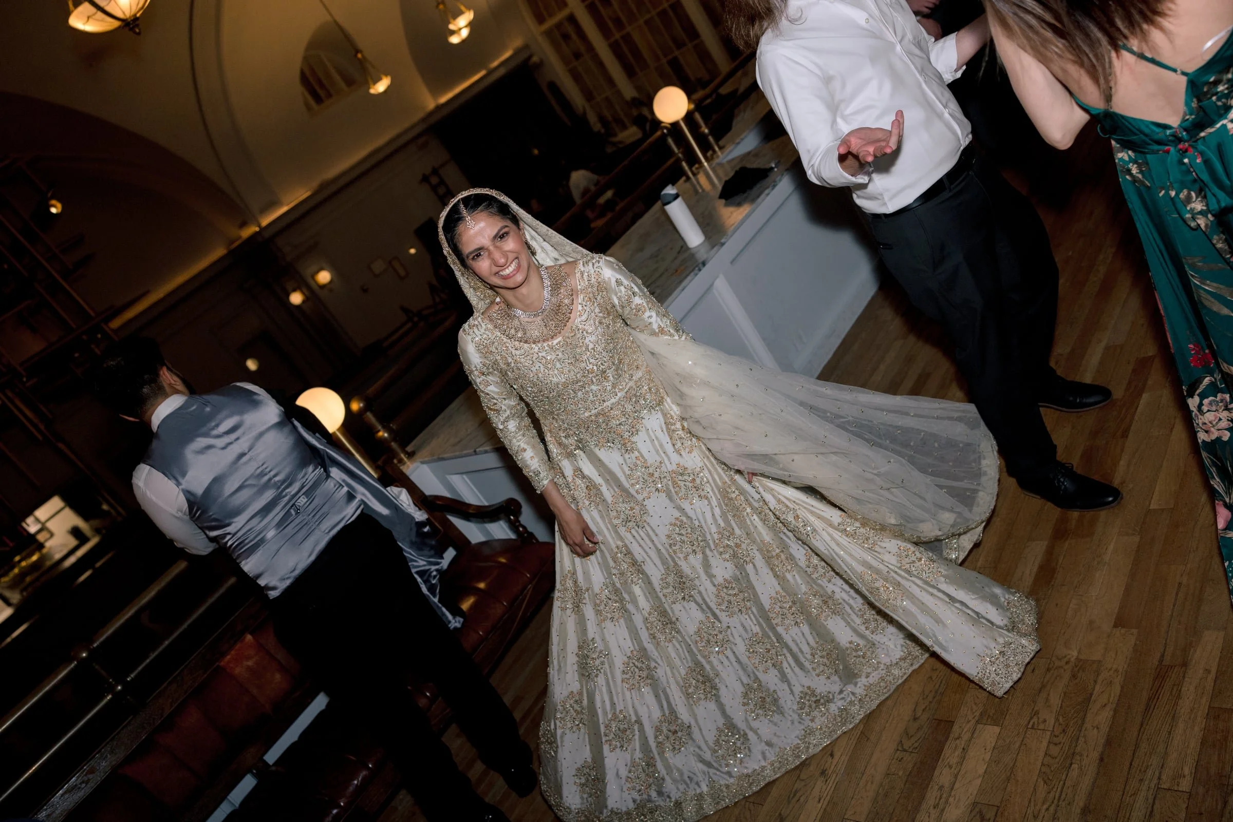 A woman wearing a gold and cream-colored ornate traditional dress with a embroidered veil and jewelry, smiling at the camera during a celebration or wedding event.