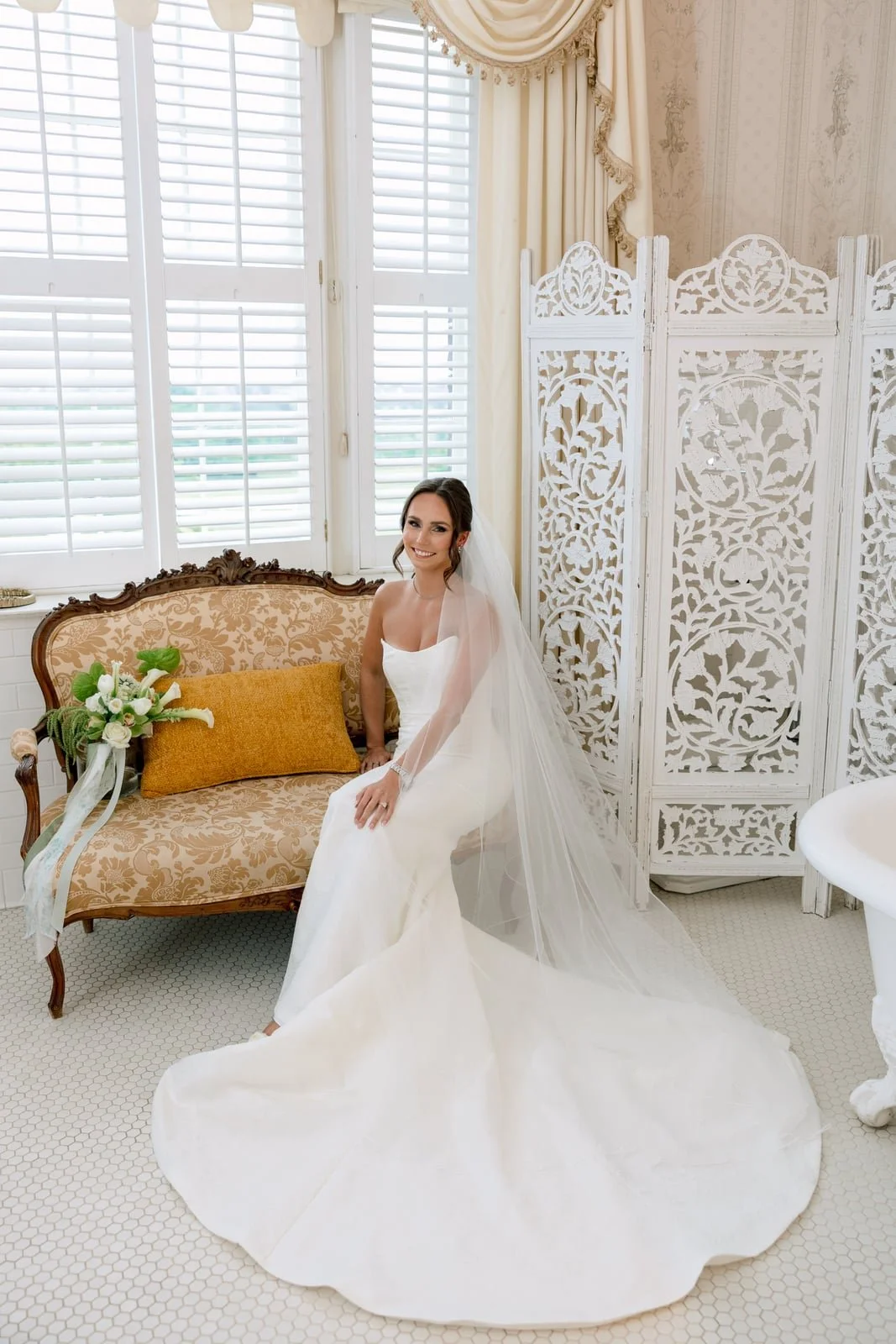 A bride in a white wedding dress sitting on a vintage sofa with floral upholstery, a bouquet of white flowers with greenery beside her, and a decorative folding screen behind her in a bright, elegantly decorated room with large window shutters.