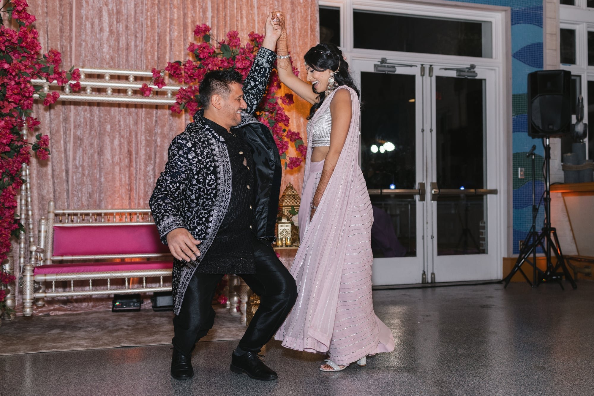 A man and a woman dancing happily at a celebration, with the woman dressed in a traditional pink outfit and the man in a black embroidered jacket, surrounded by festive decor with pink flowers.