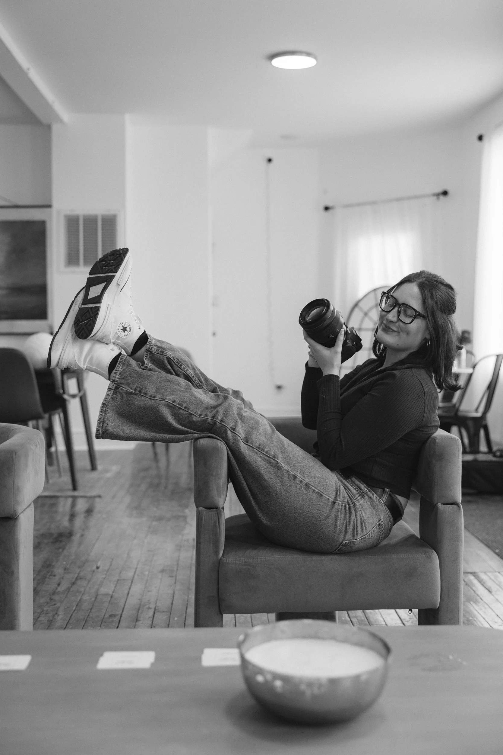A woman with glasses and long hair sitting on a chair with her legs up on another chair, holding a camera, in a living room with wooden floors and white walls.