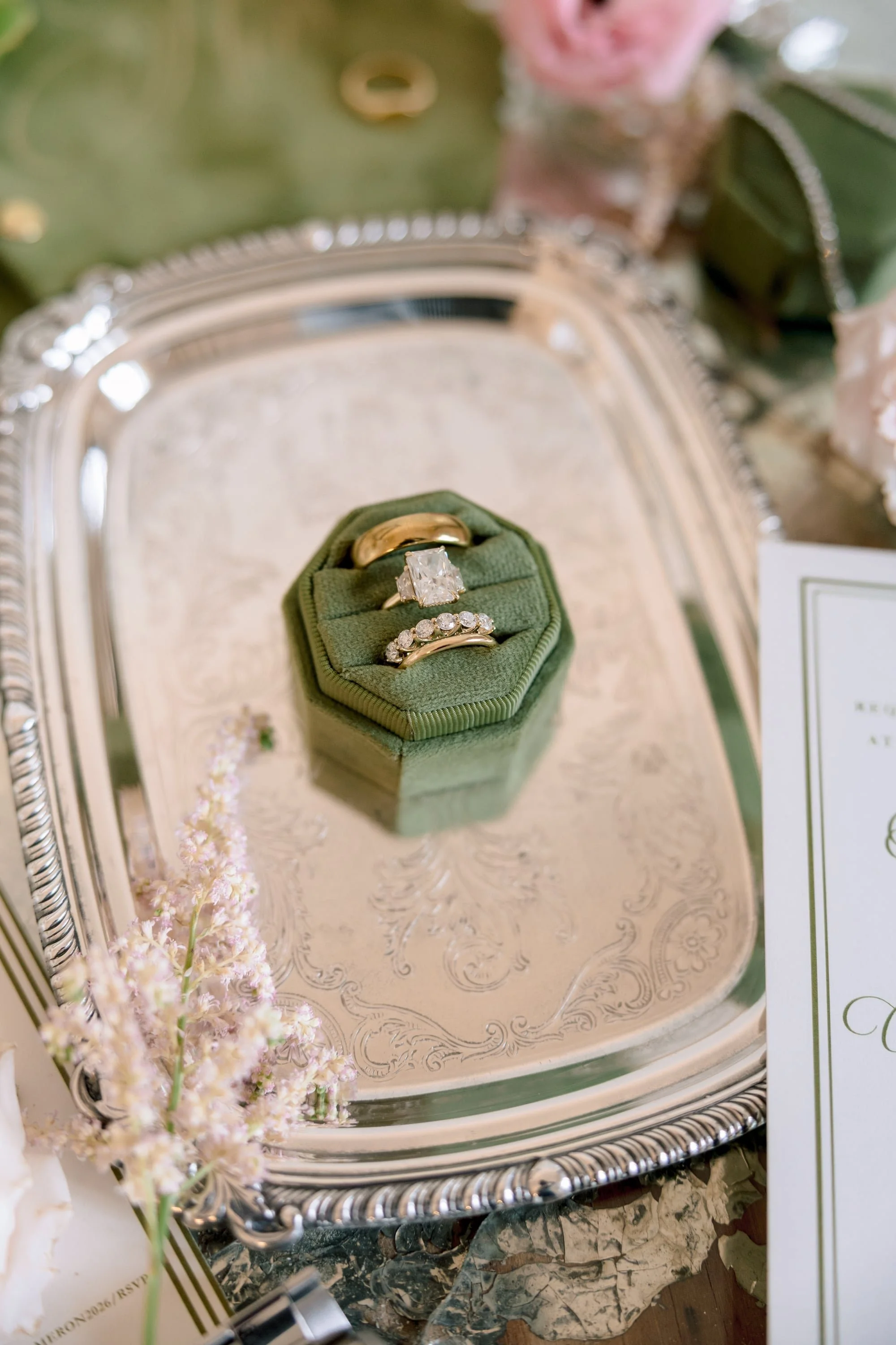 Close-up of wedding rings on a green ring box on a silver tray, with pink flowers in the background.