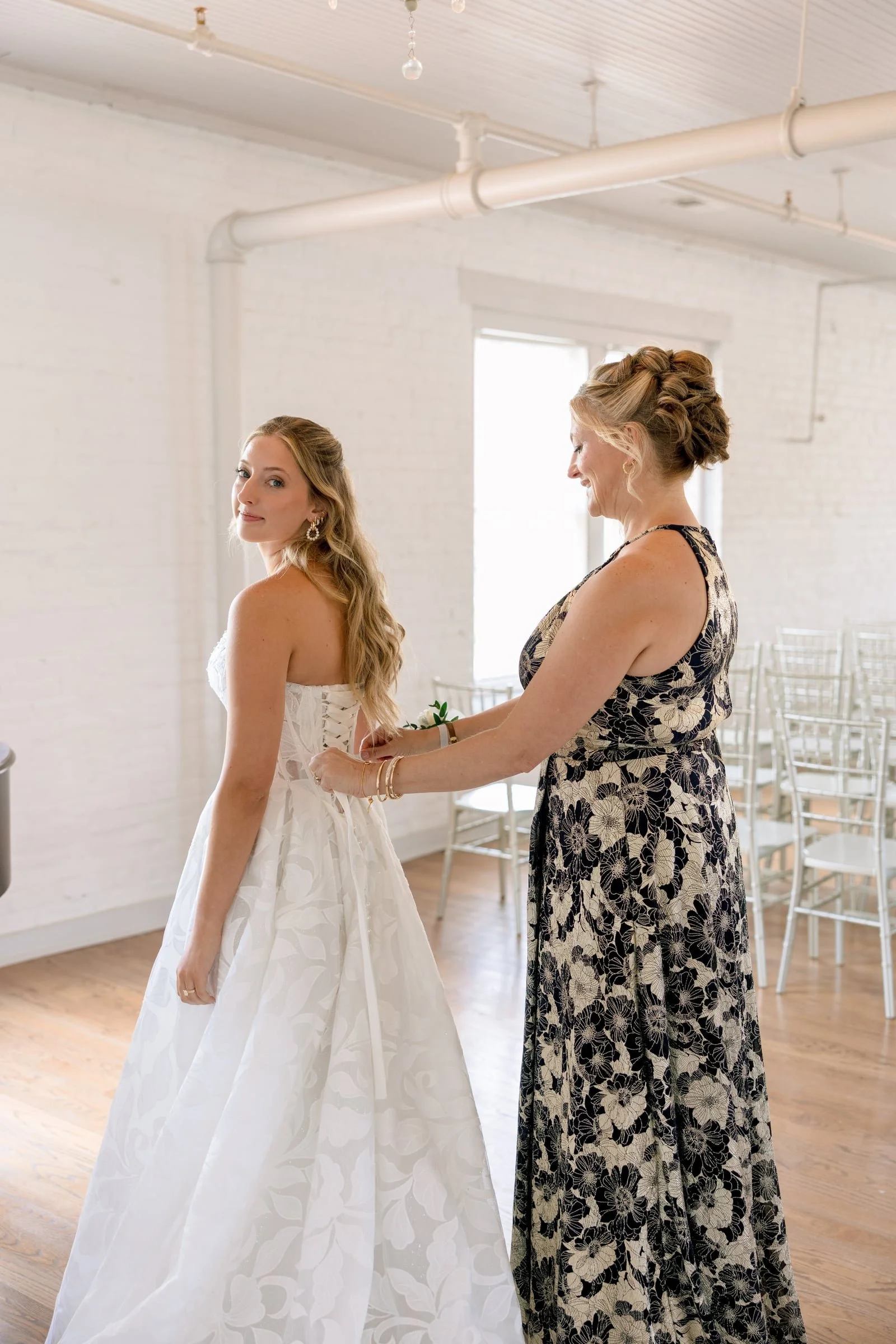 A bride in a white wedding gown standing in a room with white brick walls, while an older woman in a floral dress adjusts her dress. The room has wooden floors and white chairs, with large windows letting in natural light.