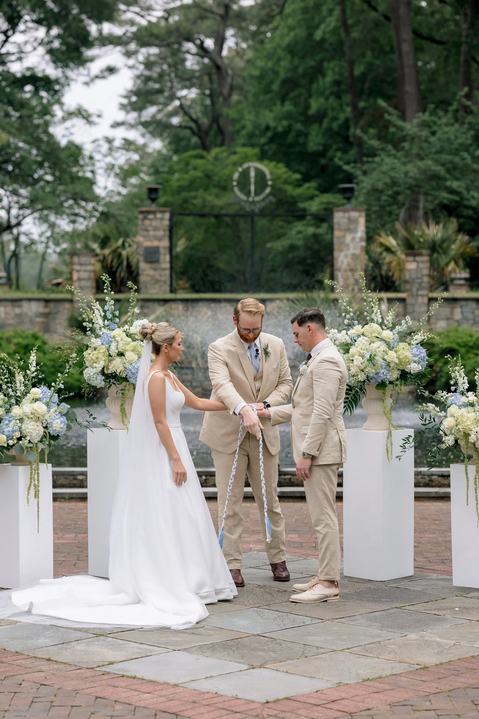 A wedding ceremony outdoors featuring a bride and groom holding hands with an officiant, surrounded by floral arrangements, with trees and a wrought-iron gate in the background.