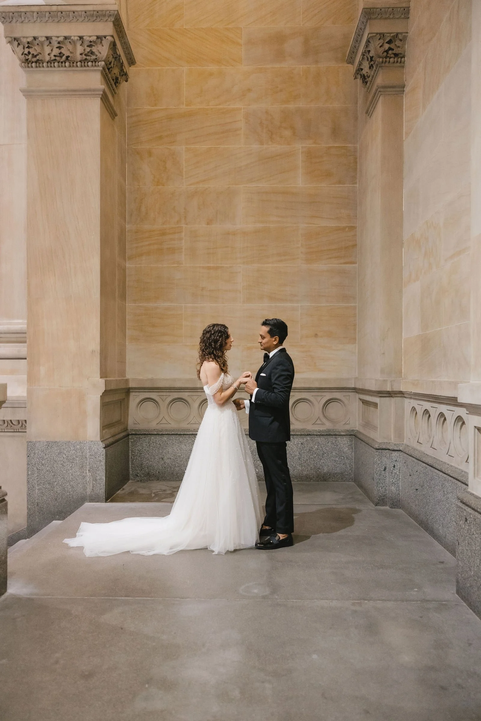 A bride and groom stand facing each other in a large, ornate hall with beige stone walls and columns. The bride wears a white gown with a long train, and the groom is dressed in a black tuxedo. They are holding hands and gazing into each other's eyes in a romantic moment.