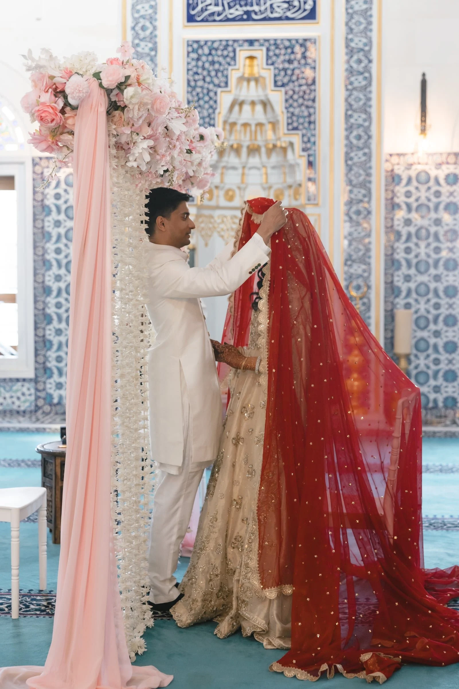 A groom in a white suit is taking off a red veil from a bride dressed in a traditional gold and beige bridal outfit with embroidery and embellishments inside a decorated venue with blue and gold motifs and floral arrangements.