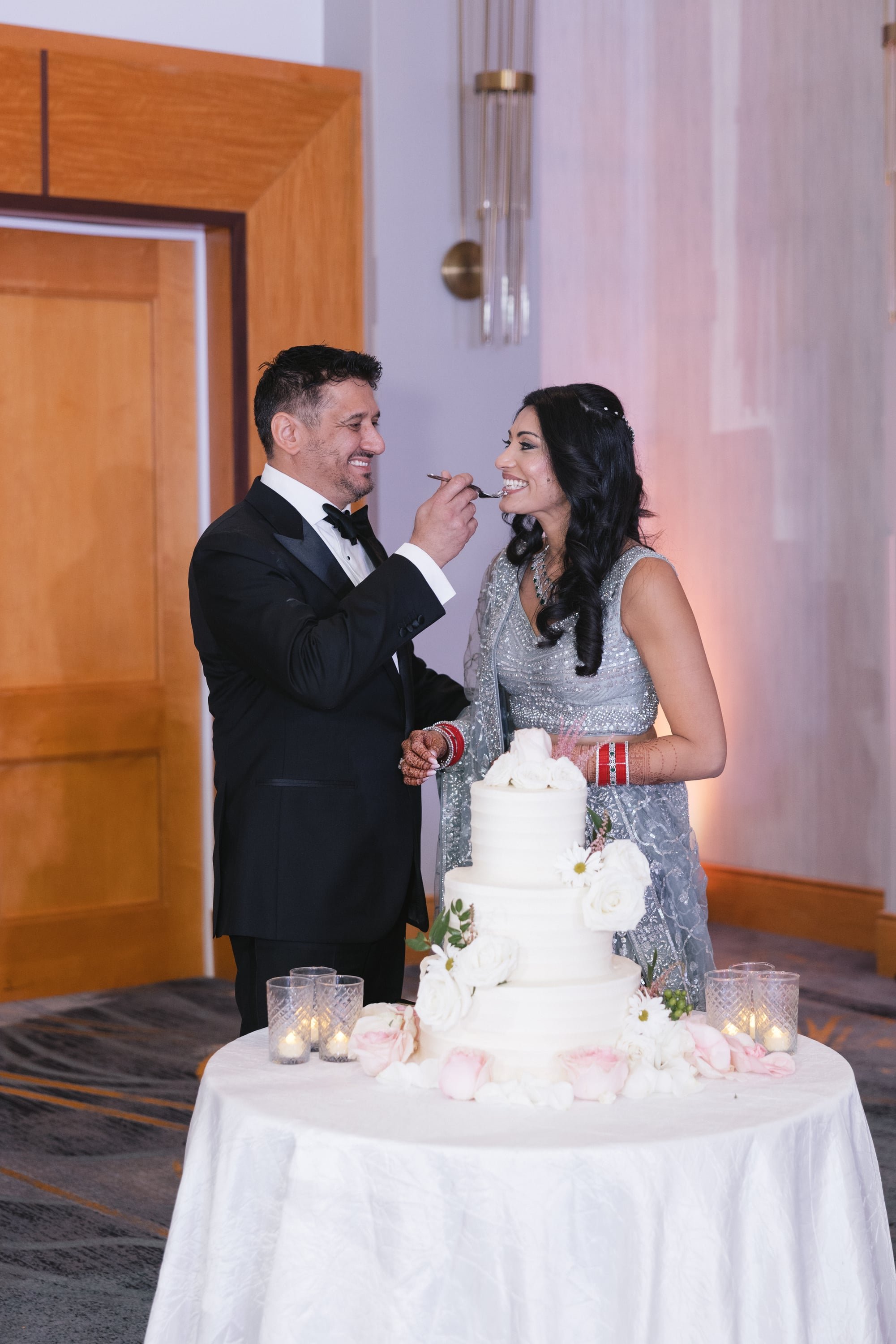 A woman in an ornate silver dress and a man in a tuxedo are at a celebration table with a large white cake decorated with white and pink flowers. The man is feeding the woman a bite of cake, and they are smiling at each other.