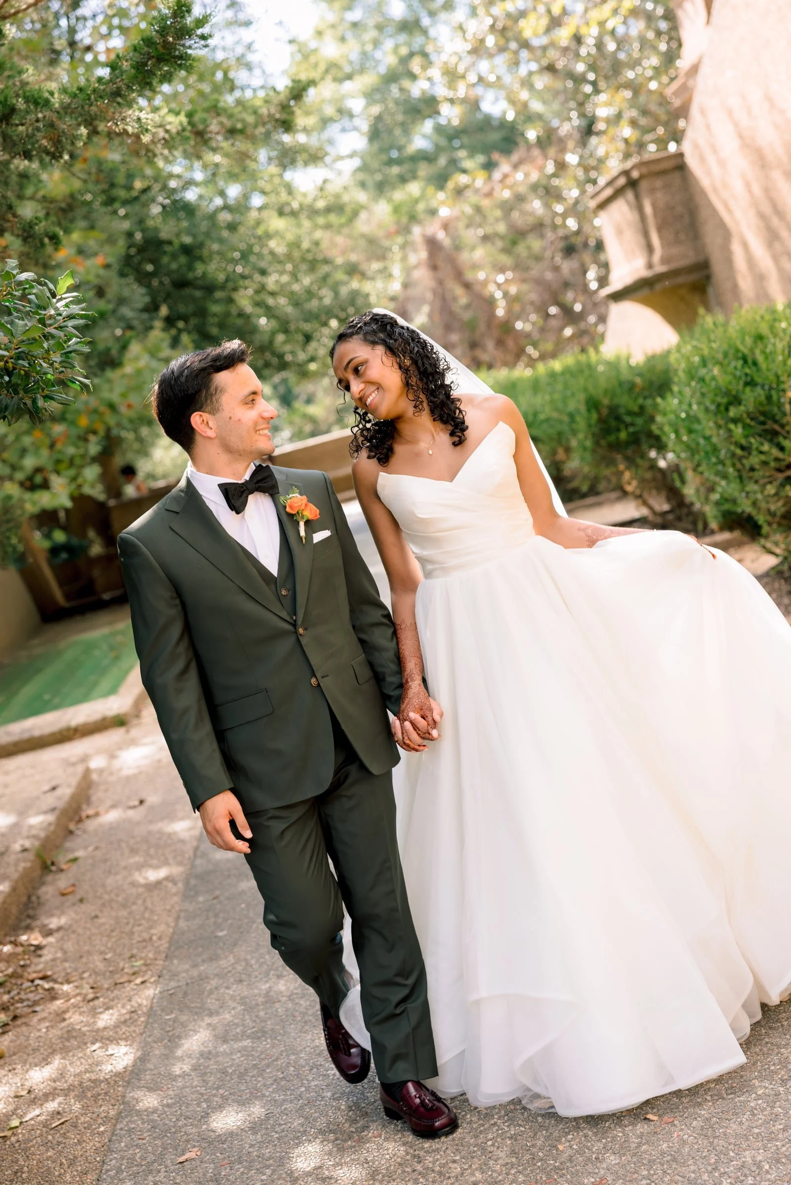 A bride and groom walking hand in hand outdoors on a pathway with trees and greenery around them. The bride is wearing a white wedding gown and veil, and the groom is dressed in a dark suit with a bowtie. They are smiling and looking at each other.