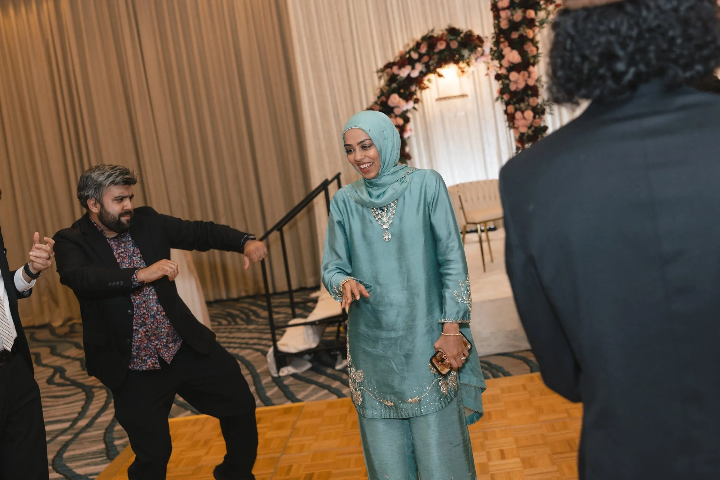 A woman wearing a turquoise dress and headscarf smiling and dancing at a social event, with a floral arch in the background.