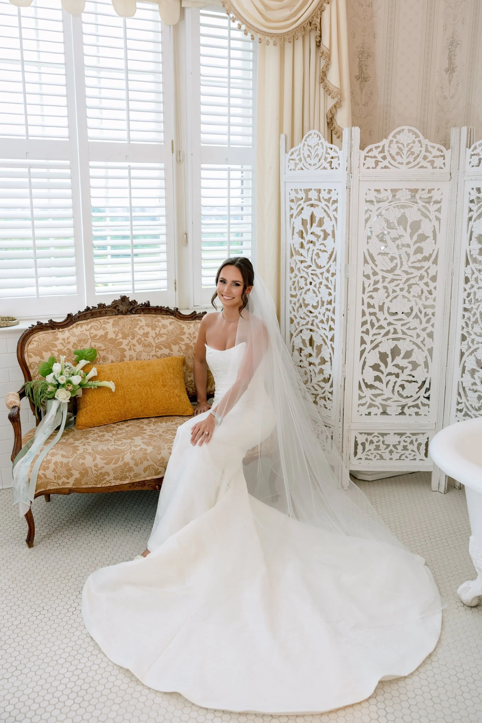 A bride in a white wedding dress sitting on a vintage sofa with floral upholstery, a bouquet of white flowers with greenery beside her, and a decorative folding screen behind her in a bright, elegantly decorated room with large window shutters.