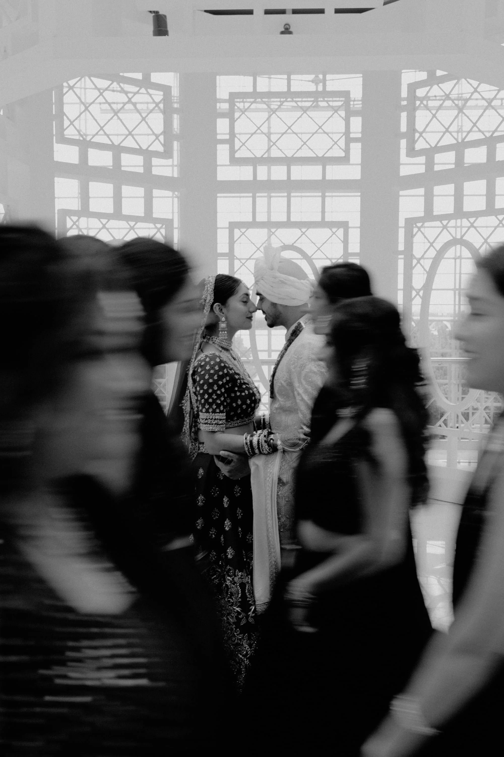 A black-and-white photo captures a couple in traditional Indian wedding attire, sharing a moment with their foreheads touching, while people walk by in a busy indoor setting with large, ornate windows in the background.