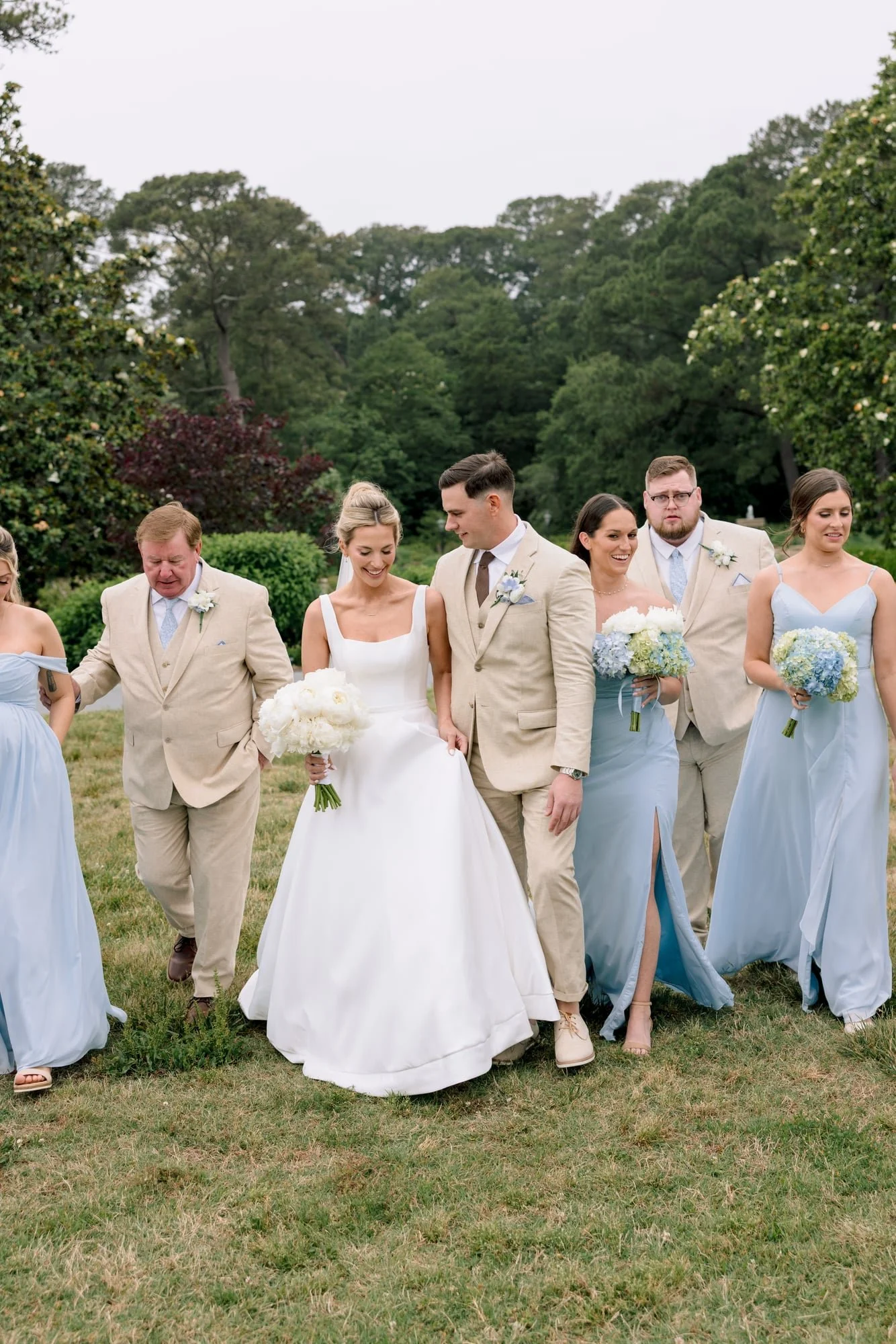 Group of people dressed in wedding attire walking outdoors on a grassy field surrounded by trees.