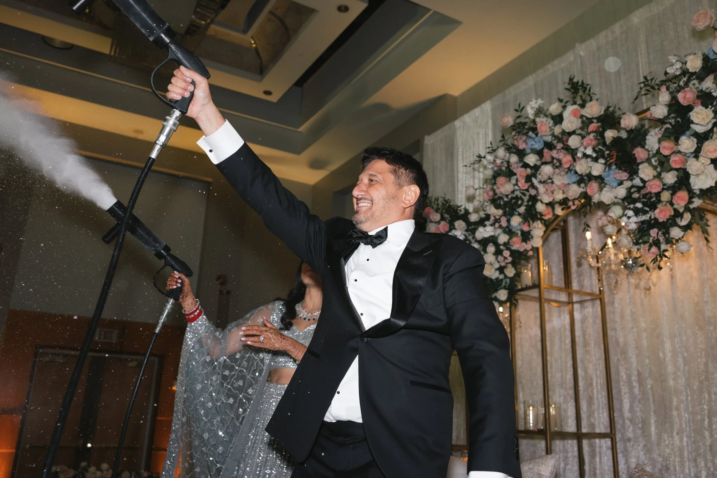 A man and a woman dancing at a wedding reception. The man is wearing a black tuxedo and the woman is wearing a sparkly silver dress. They are holding a fog machine. The background features floral decorations and elegant lighting.