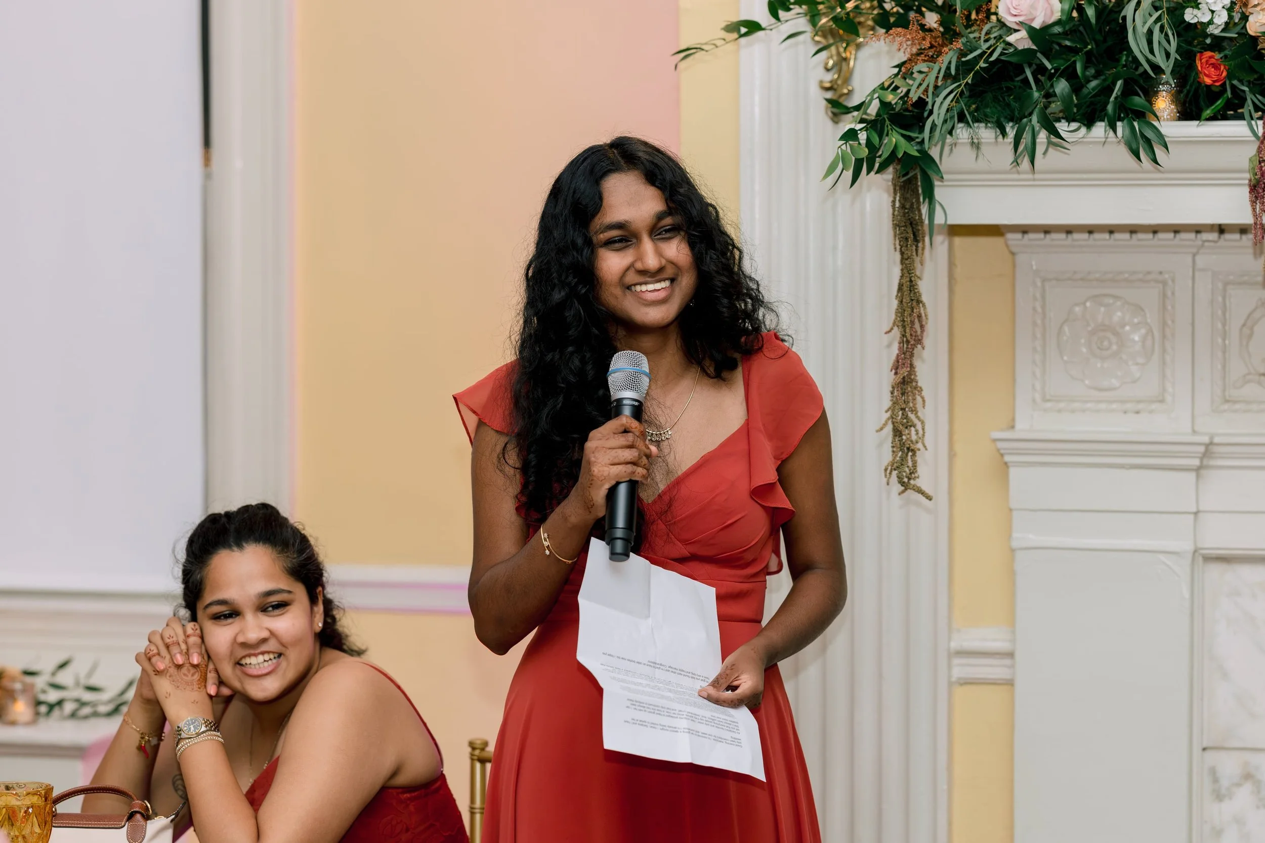 A woman in a red dress holding a microphone and a piece of paper, smiling at an event. Two women are partially visible, one sitting with her hand on her face, at a decorated indoor venue with floral arrangements.