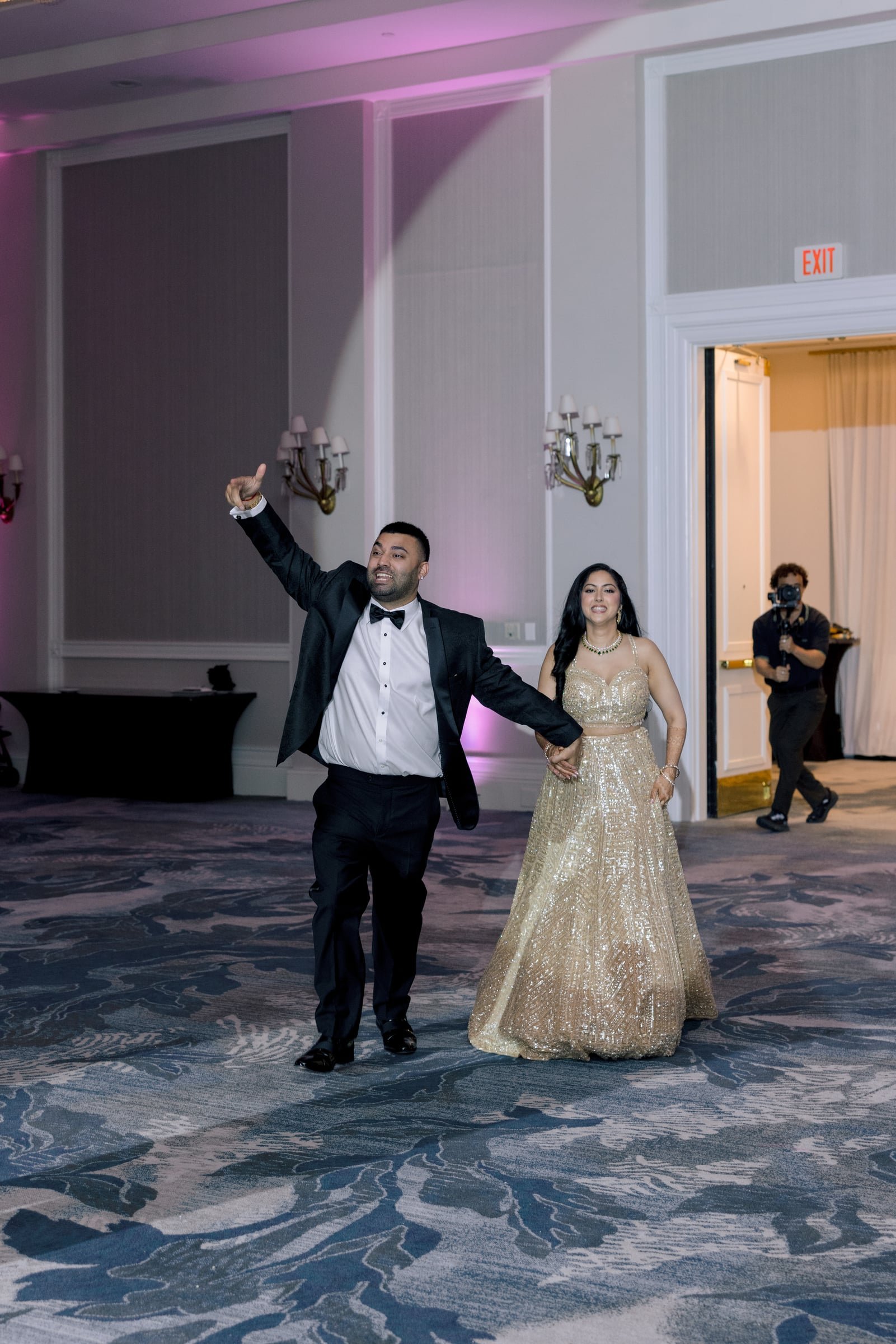 A couple dancing at a formal event, with the man in a tuxedo and the woman in a gold, sequined gown. An individual with a camera is in the background.