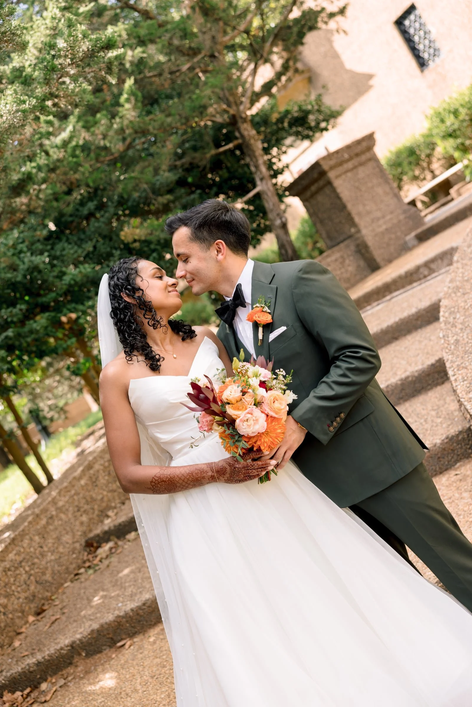 A bride and groom sharing a tender moment outdoors, the bride holding a bouquet of pink and orange roses, surrounded by trees and stone steps.