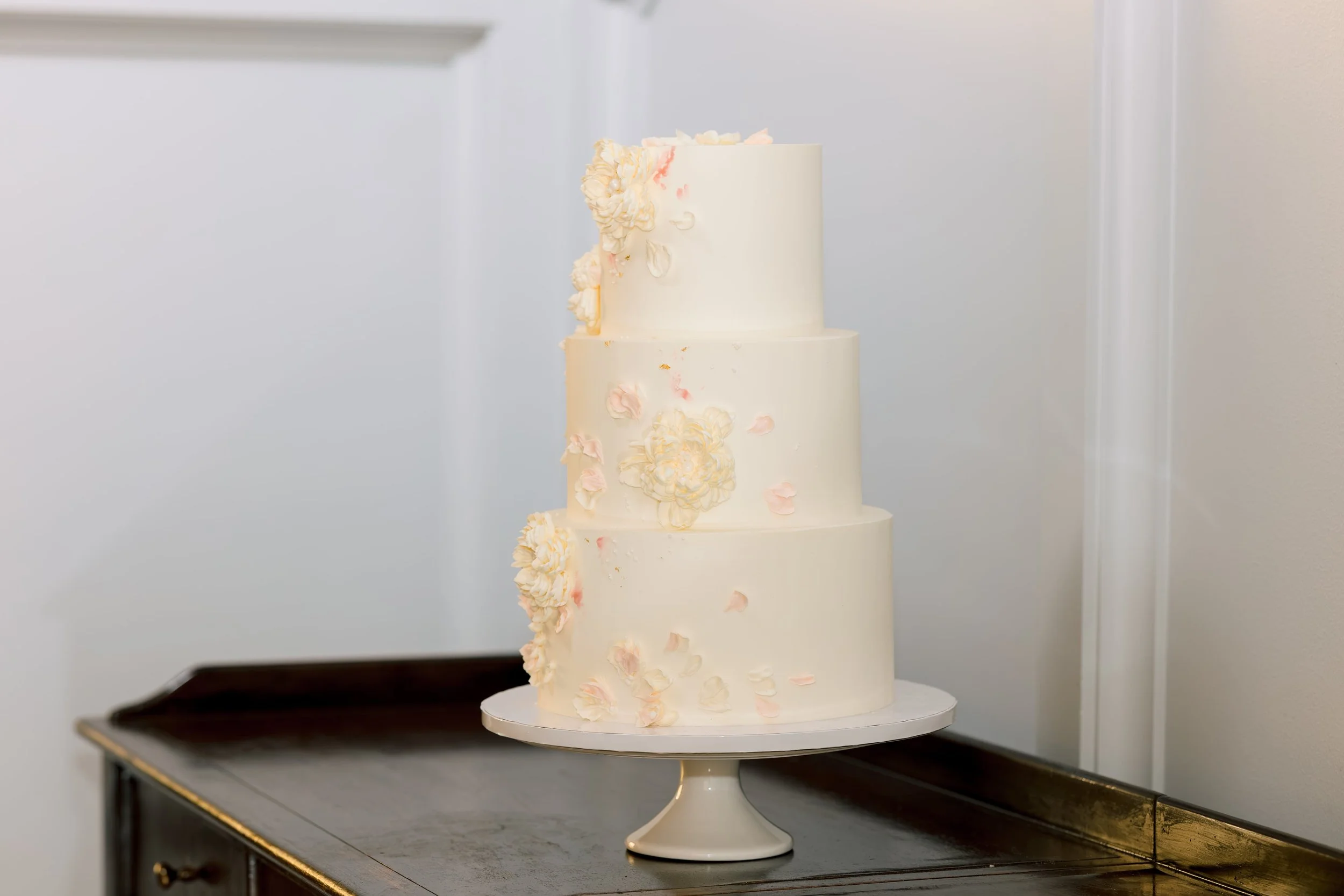 A tall, white, multi-tiered wedding cake decorated with soft pink and white flower embellishments on a white cake stand.