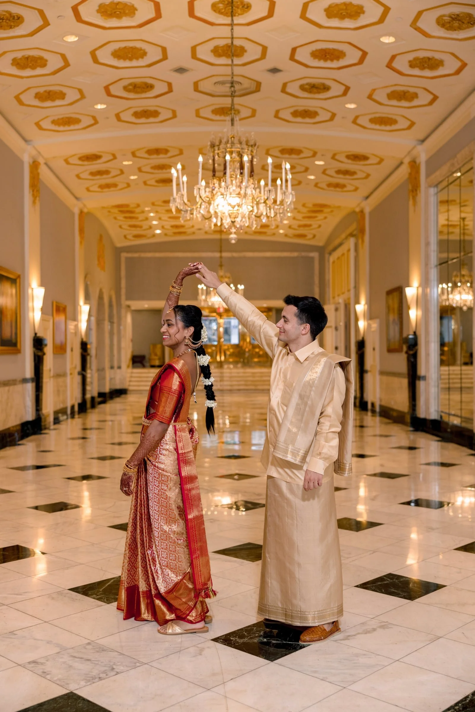 A couple dressed in traditional Indian attire dancing in a grand hallway with ornate ceiling and chandeliers.