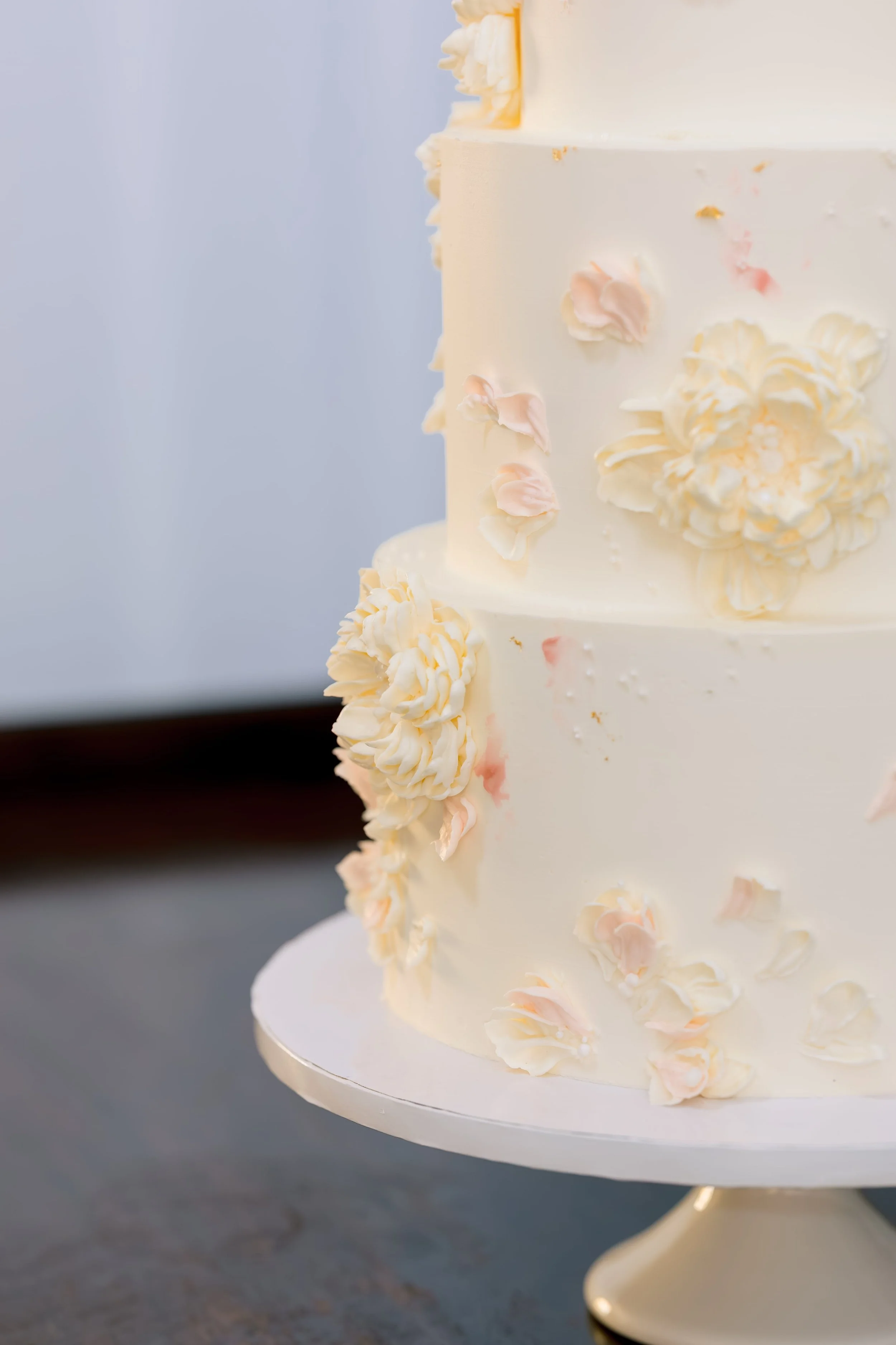 A white, multi-tiered wedding cake decorated with pink and white flower petals and cream-colored rosettes, sitting on a white cake stand.