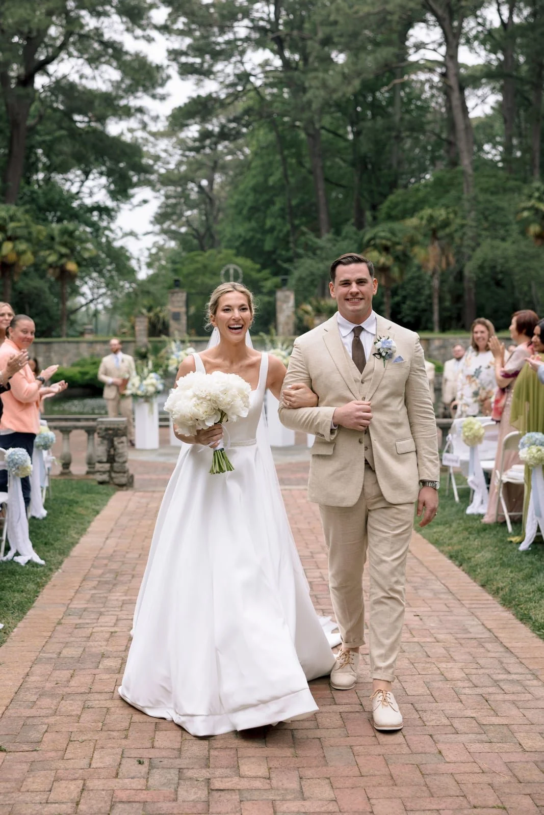 Bride in a white gown holding flowers and smiling, walking arm-in-arm with groom in a beige suit, during outdoor wedding ceremony with guests applauding, in a lush garden setting.