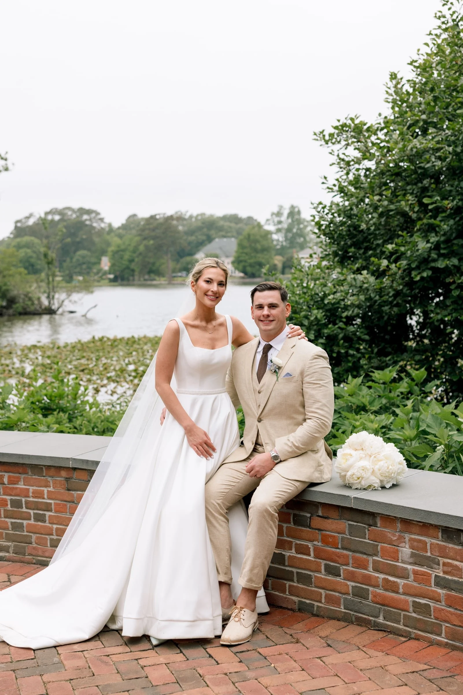 A newlywed couple smiling outdoors by a river, with the bride in a white wedding dress and the groom in a beige suit, sitting on a brick ledge with a bouquet of white flowers nearby, surrounded by green trees and overcast sky.