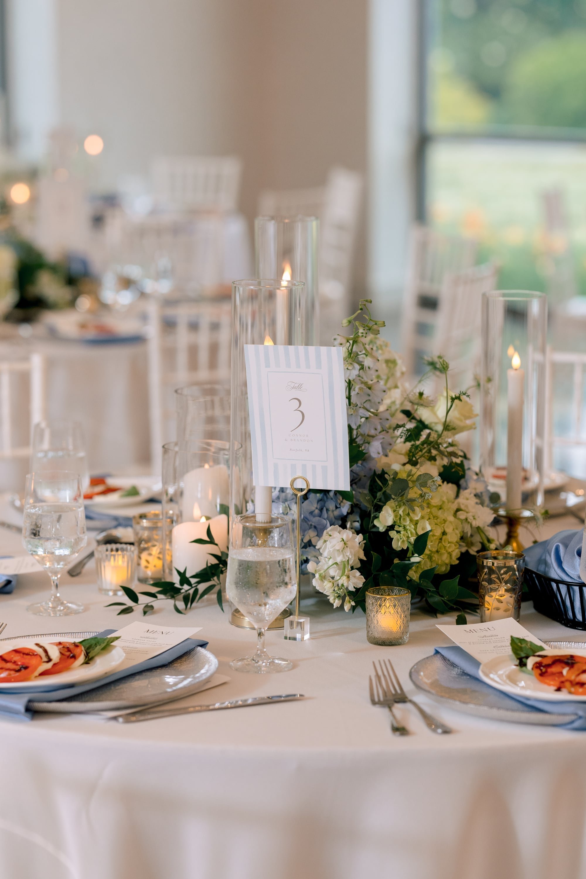 Elegant table setting with white tablecloth, floral centerpiece, candles, water glasses, plates with appetizers, and table number with floral decoration in a bright, airy room with large windows.