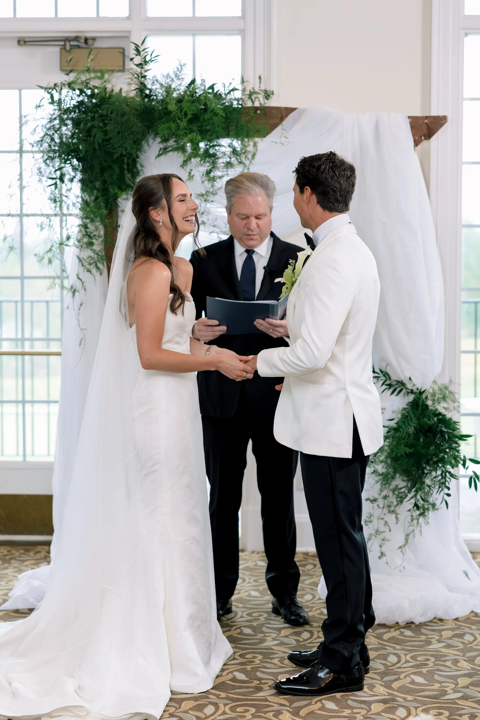 A bride and groom holding hands during their wedding ceremony, with an officiant reading from a book.