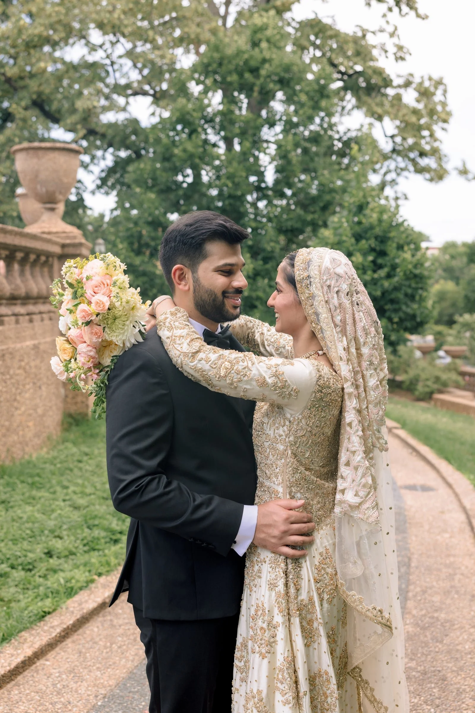 A bride and groom are embracing outdoors during a wedding, with the bride wearing a traditional white and gold embroidered dress and a headscarf, holding a bouquet of pink and white roses, while the groom is dressed in a black tuxedo with a bow tie, 