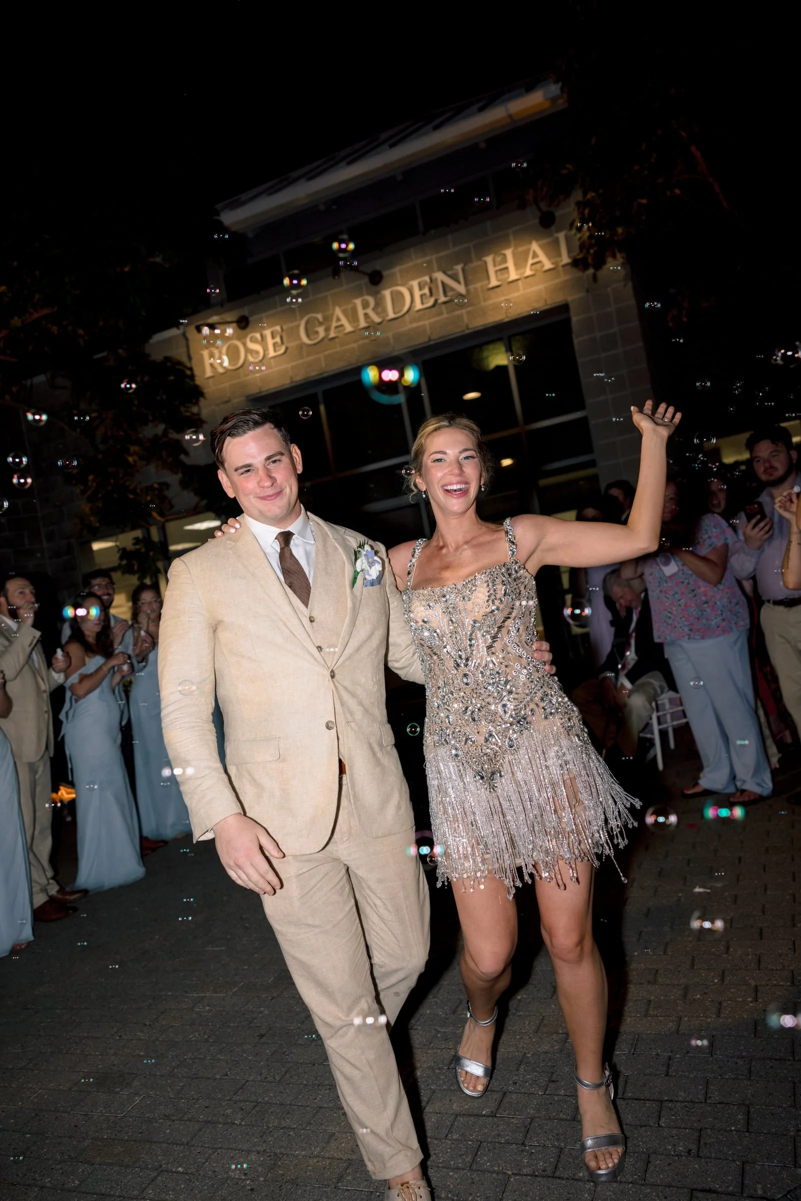 A couple dancing at an event outside a building with a sign that reads 'Rose Garden Hall.' The man wears a beige suit with a white shirt and brown tie, and the woman wears a silver, beaded dress with fringe, both smiling.