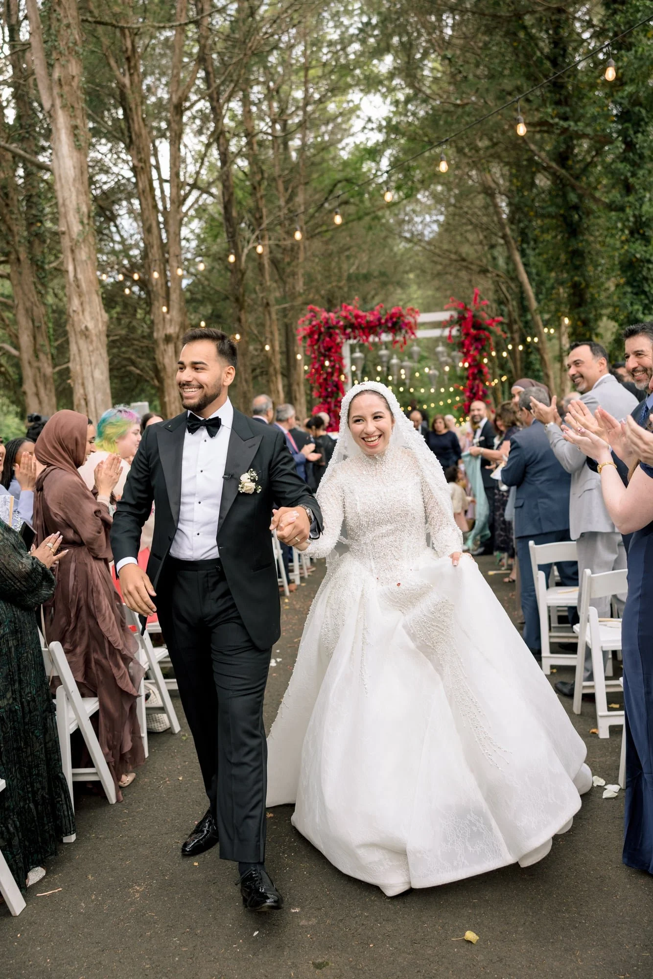 A bride and groom walking down the aisle at an outdoor wedding ceremony, surrounded by friends and family, with trees and string lights in the background.