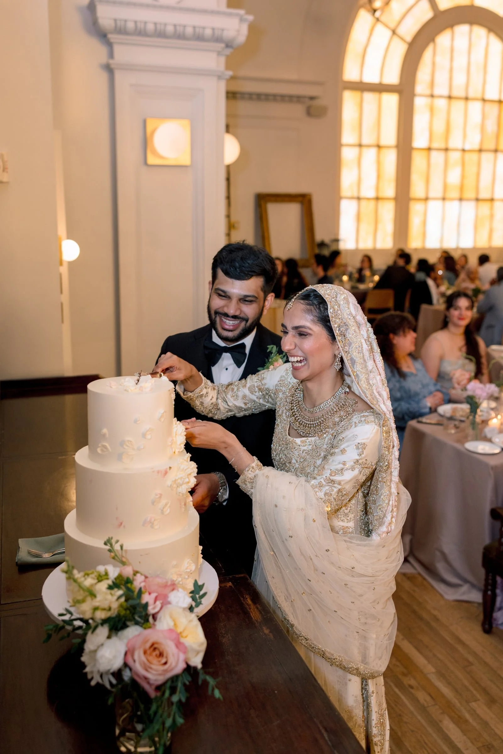 A bride and groom are cutting a wedding cake together at their reception. The bride is dressed in a traditional, ornate wedding dress with a head covering, and the groom is wearing a black tuxedo. The cake is three-tiered, decorated with flowers, and