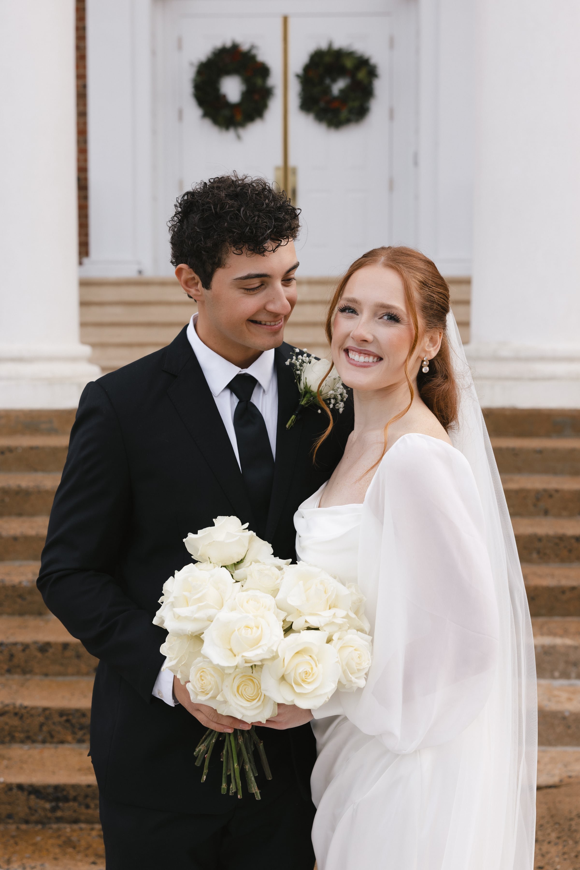 A smiling bride with red hair and a groom with curly black hair standing together on the steps of a church, holding a large bouquet of white roses.