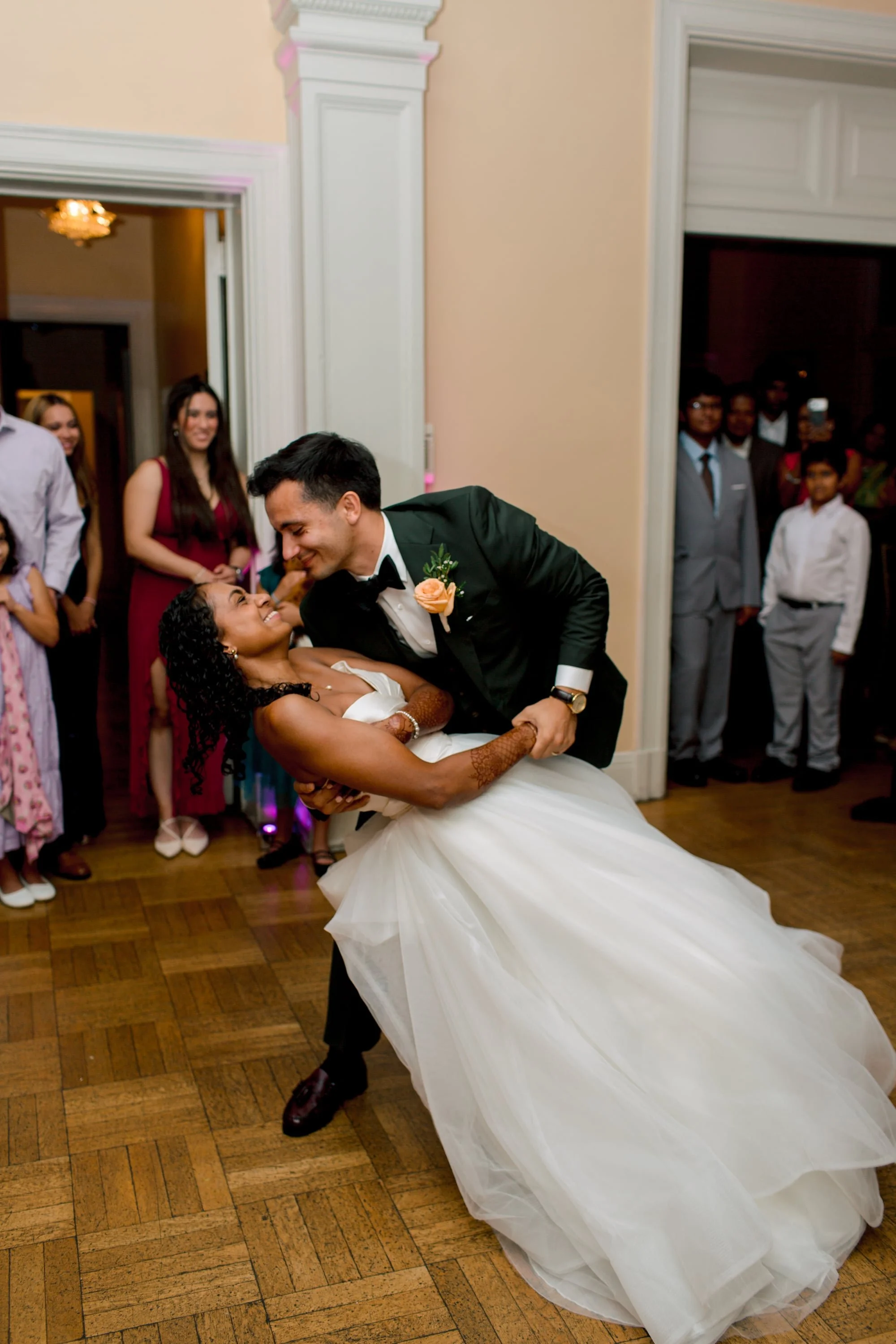 A groom in a tuxedo with a yellow flower boutonniere dances with a bride in a white wedding dress at a wedding reception, with guests watching in the background.