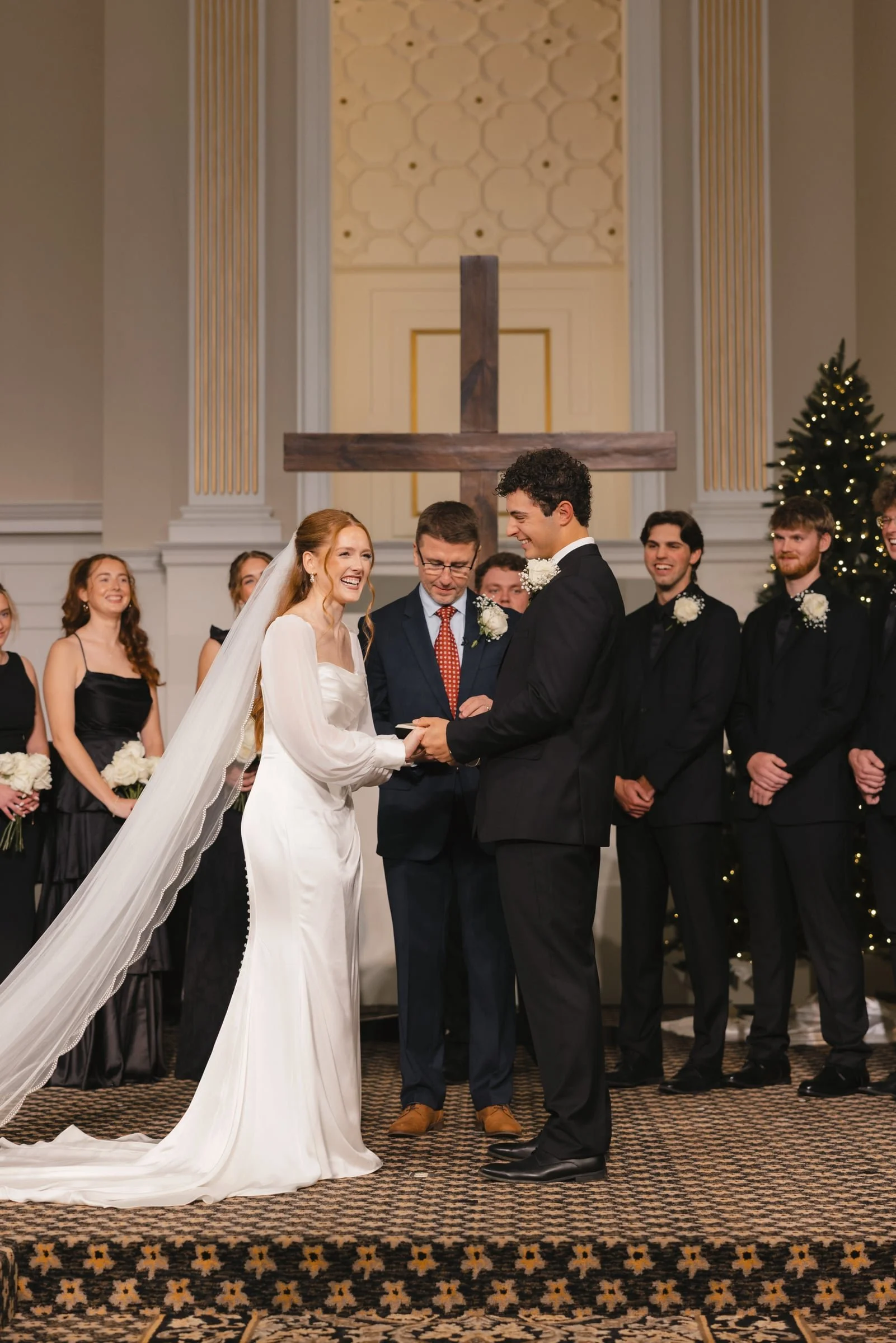 A wedding ceremony taking place inside a church with a cross in the background. The bride and groom are smiling at each other, holding hands. The bride has long red hair and is wearing a white gown with a long train. The groom has dark curly hair and