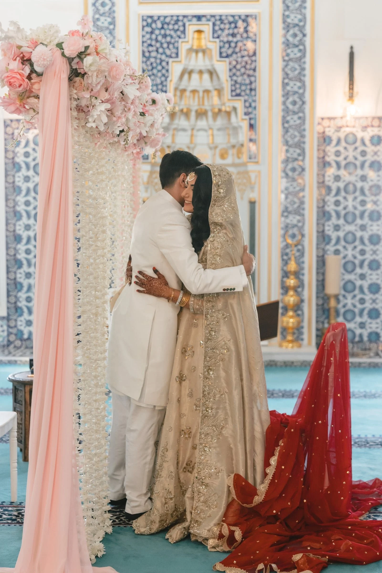 A newlywed couple embrace during their wedding ceremony inside a decorated hall with ornate blue and gold patterns. The bride is dressed in a beige embroidered gown with a matching headscarf, and the groom is in a white suit. There are pink and white