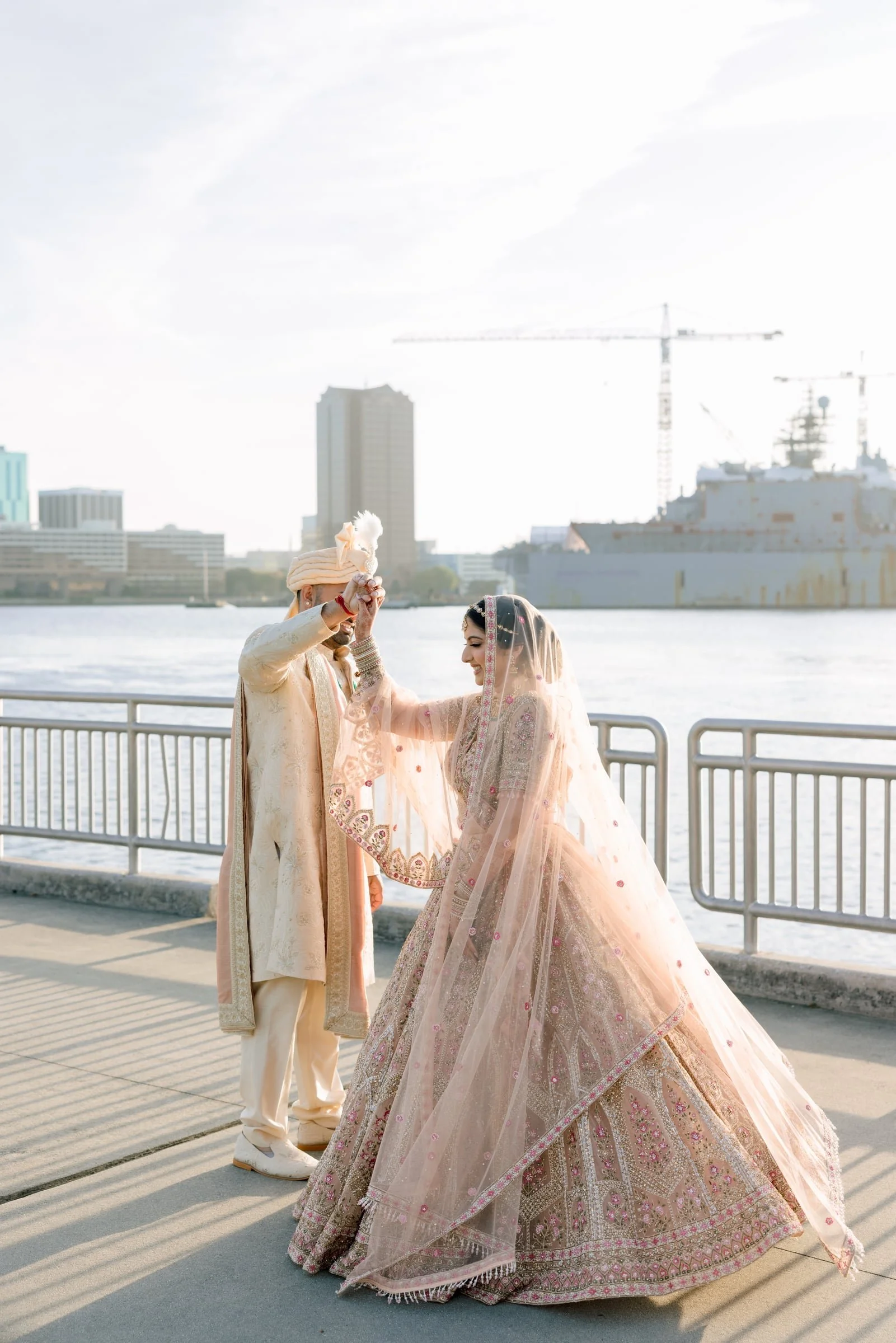 A couple dressed in traditional Indian wedding attire, standing by the water on a waterfront promenade with city buildings and construction cranes in the background. The groom is wearing a cream-colored sherwani with a turban, and the bride is in a p