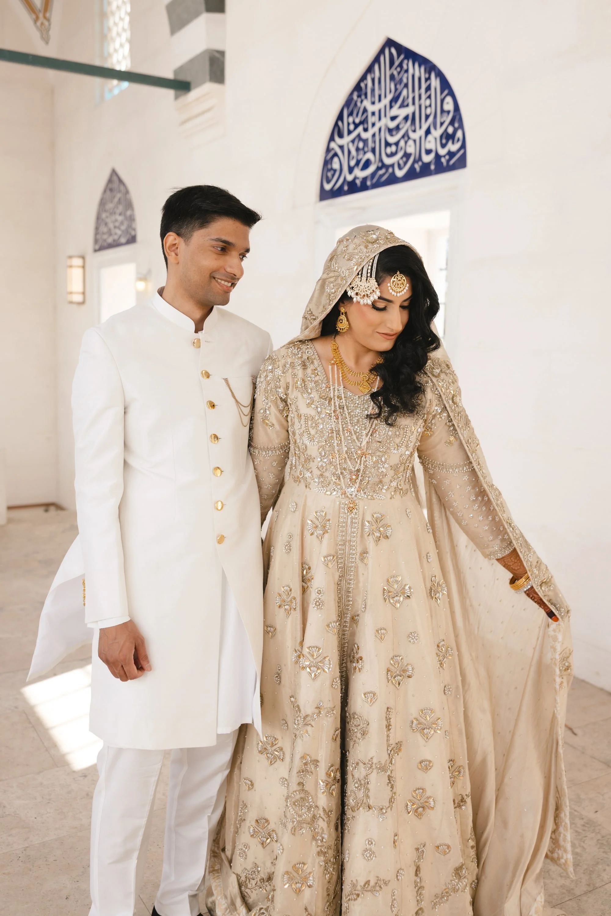 A bride and groom in traditional wedding attire standing together indoors. The bride wears a heavily embroidered cream-colored dress with gold jewelry and a head covering. The groom wears a white suit with gold buttons and a matching chain.